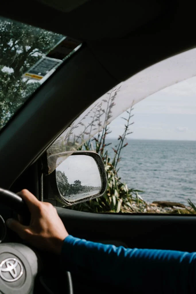 Water and rocky coastline is visible out the window of a car