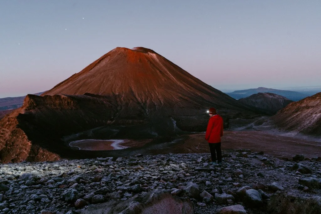 A woman stands in front of a view of Mount Ngauruhoe in Tongariro National Park, with her headtorch focused on the ground and dawn colours in the sky
