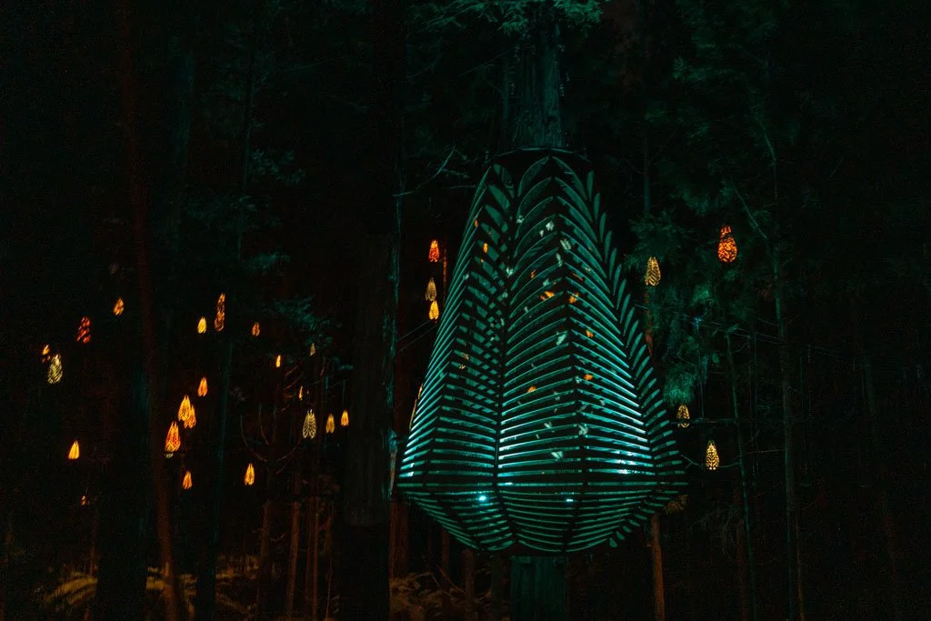 A large sculture suspended from a tree is lit up in blue light