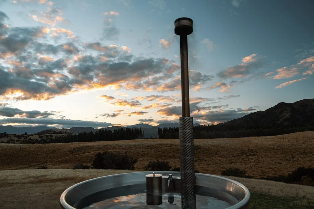 A hot tub with a chimney puffing smoke and in the background a sky filled with sunset colours