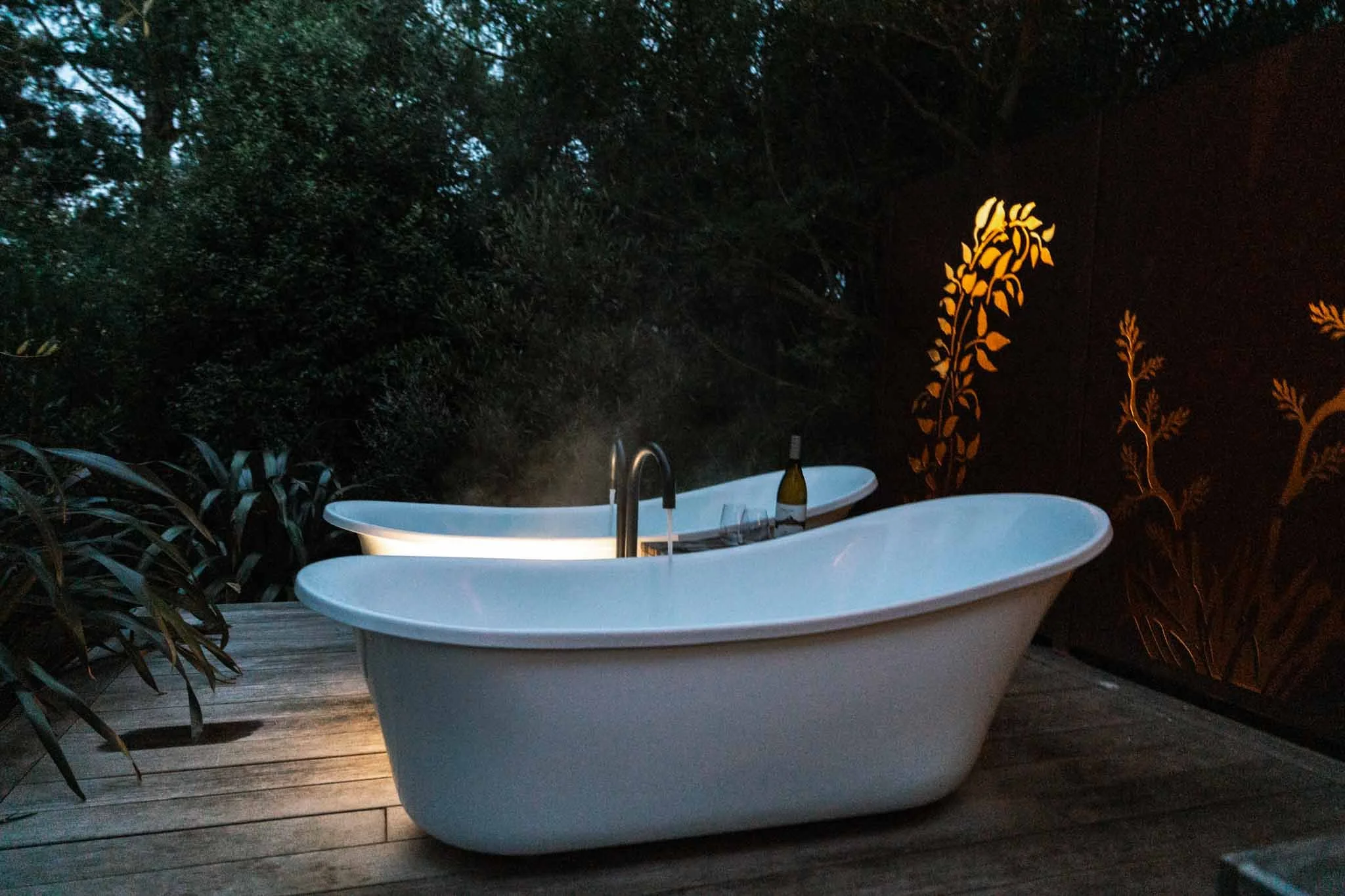 Two outdoor bathtubs on a deck with a bottle of wine in between