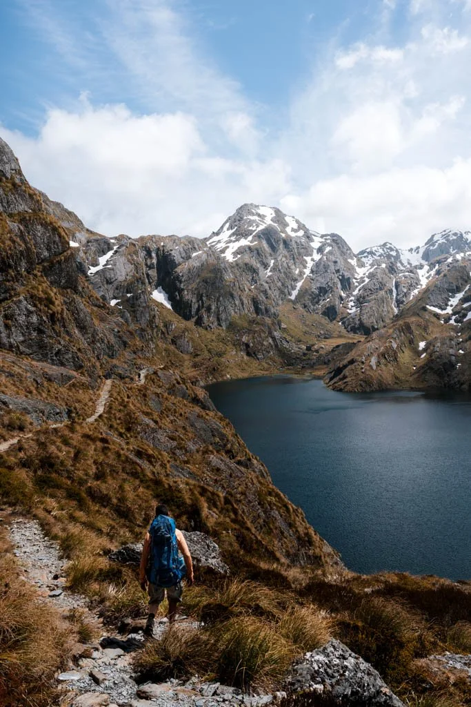 A man walks along a rocky path above a lake at Harris Saddle on the Routeburn Track