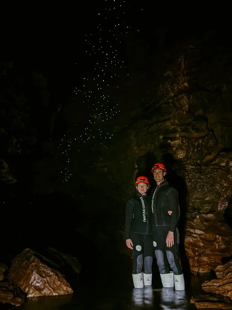 A couple wearing wetsuits and red helmets pose in from of glow worms in a cave in Waitomo