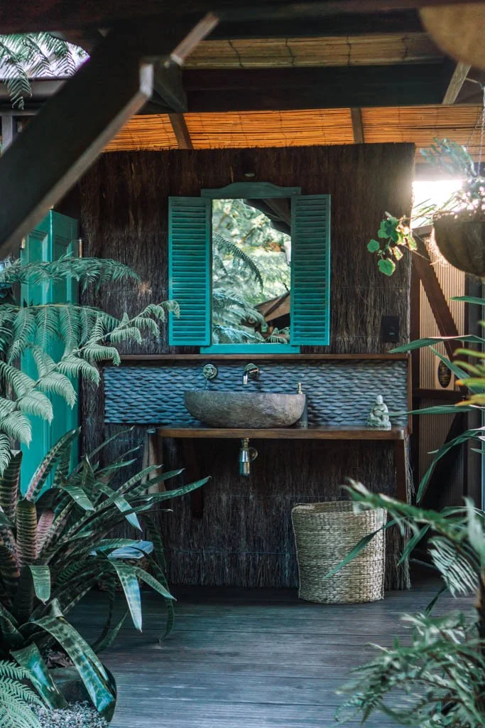 A stone sink beneath a mirror is outside, surrounded by lush ferns