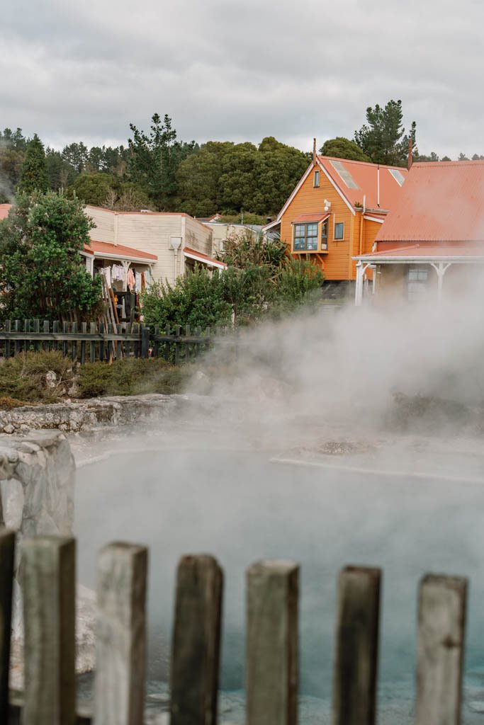 Yellow and orange cottages are visible beyond a steaming hot pool and wooden fence