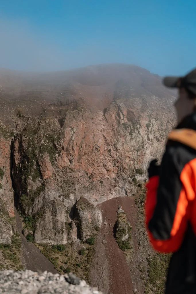 A girl wearing an orange jacket stands and looks at the crater of Mt Tarawera
