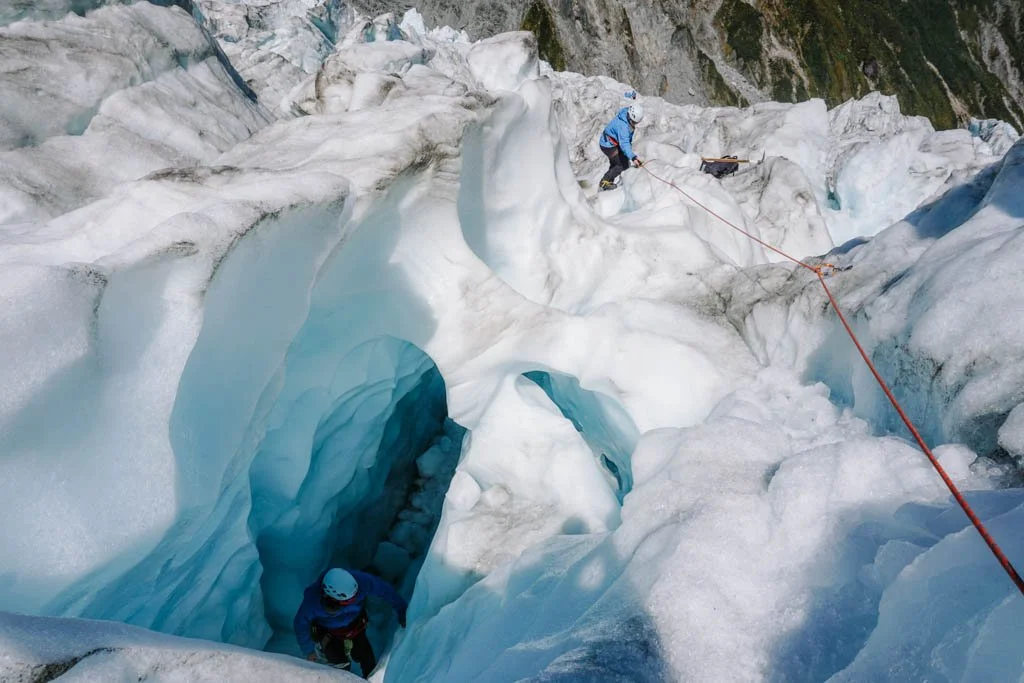 A person climbs up out of an ice cave on Franz Josef glacier, while another descends a white slope holding onto a red rope