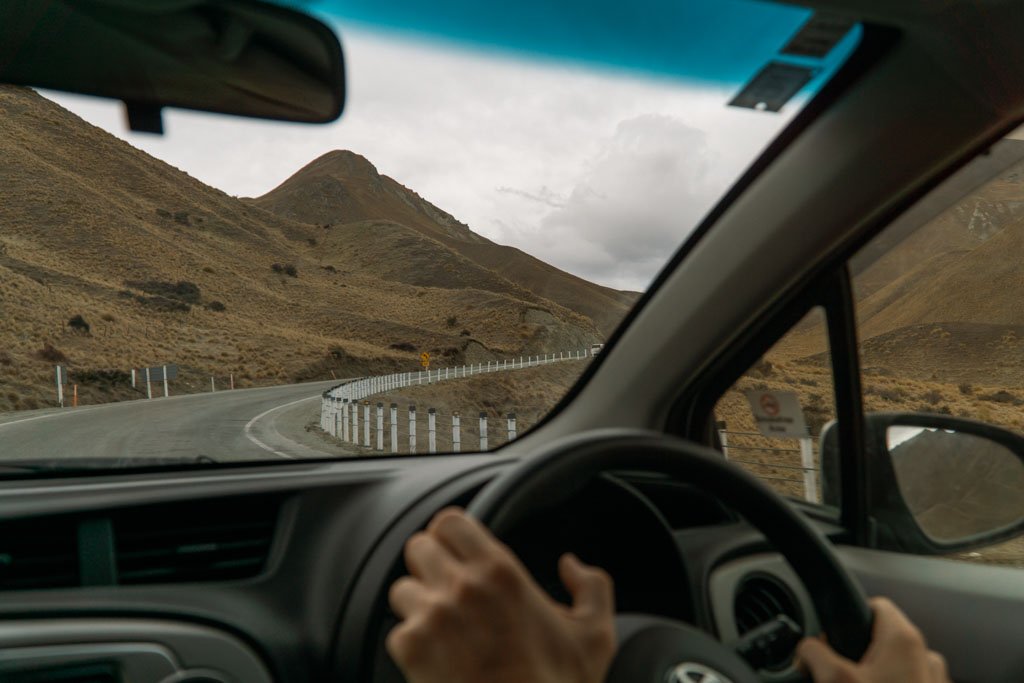 Hands on the steering wheel of a car driving through tussocked hills
