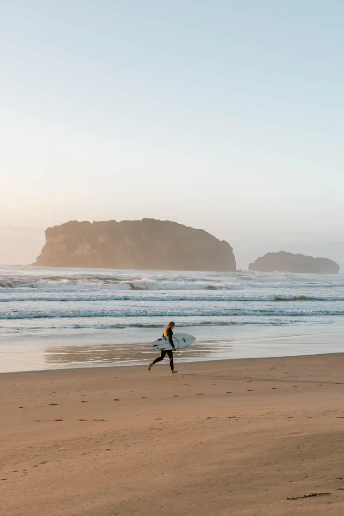 A girl holding a surfboard runs up a beach with waves and an island in the background bathed in golden morning glow