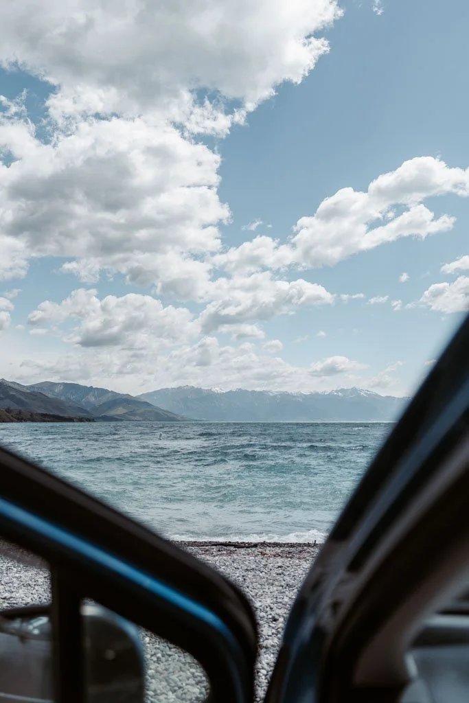A rental car is parked on a pebble beach overlooking a lake in the South Island