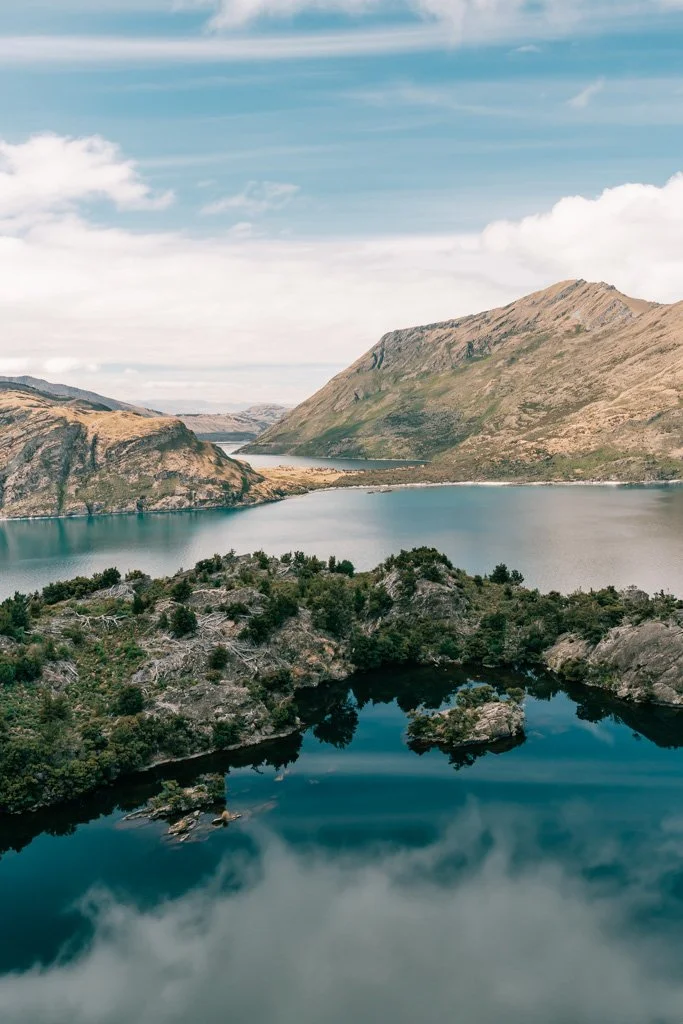 A view across a small lake surrounded by rocks, out across larger lake Wanaka, seen from the top of Mou Waho island