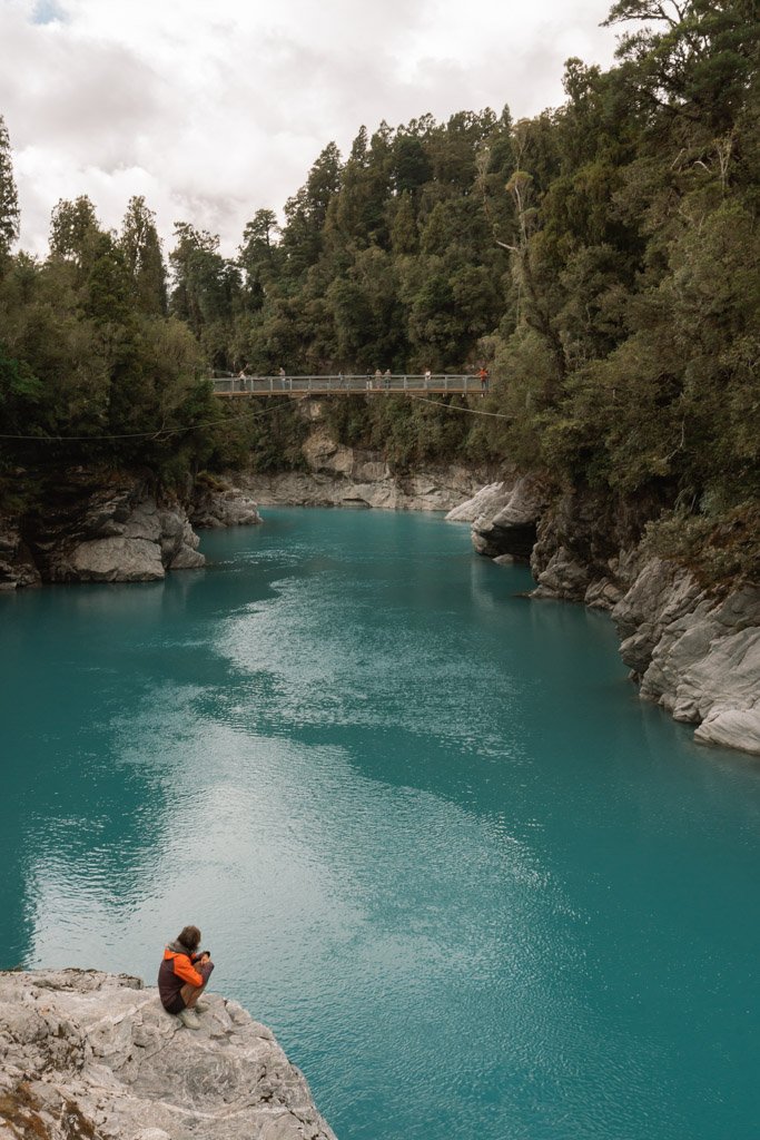 A person in an orange jacket crouches on a rock overlooking the bright blue Hokitika Gorge