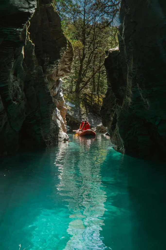 An inflatable red canoe is paddled by two people through a narrow canyon with bright blue water