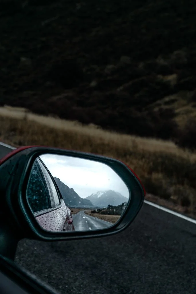 In the rear view mirror of a car, a road stretches towards mountains