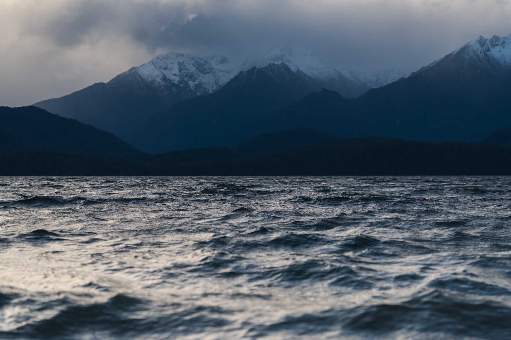 Choppy water is in the foreground and across the lake is snowy mountains draped in cloud