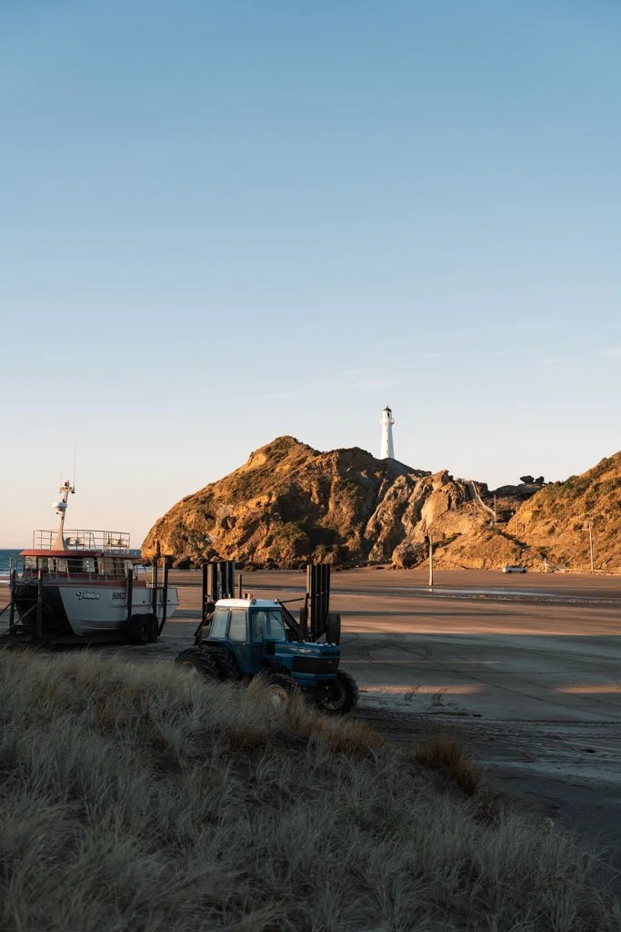 A tractor and fishing boat sit on the sand in front of a lighthouse atop a rocky point