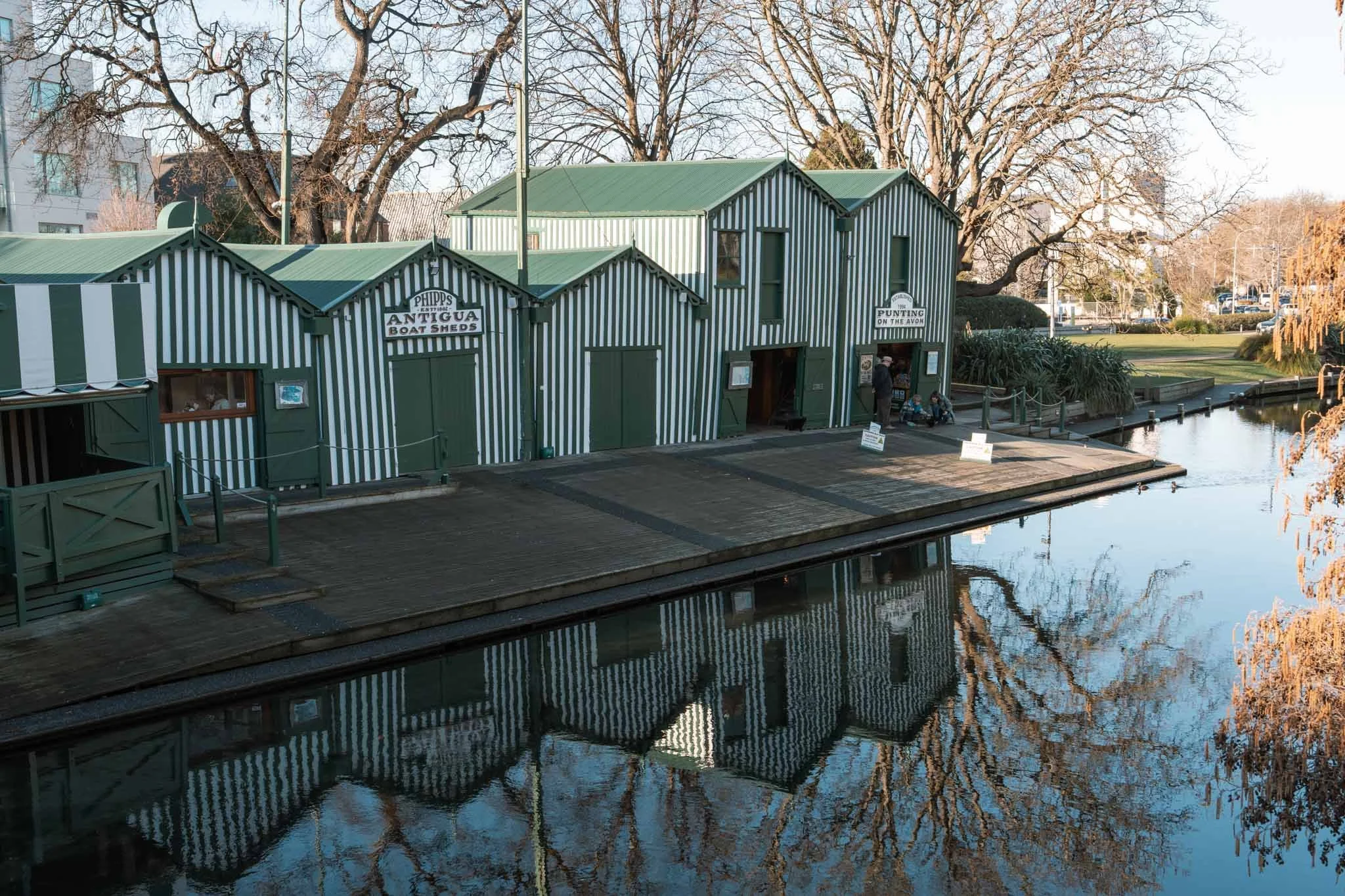 The green and white striped boat sheds in Christchurch are reflected in the still river