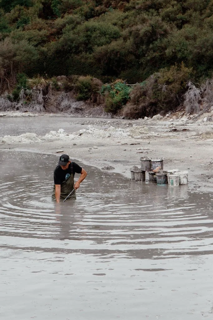 A person wearing waders stands in a mud pool and is shovelling out mud