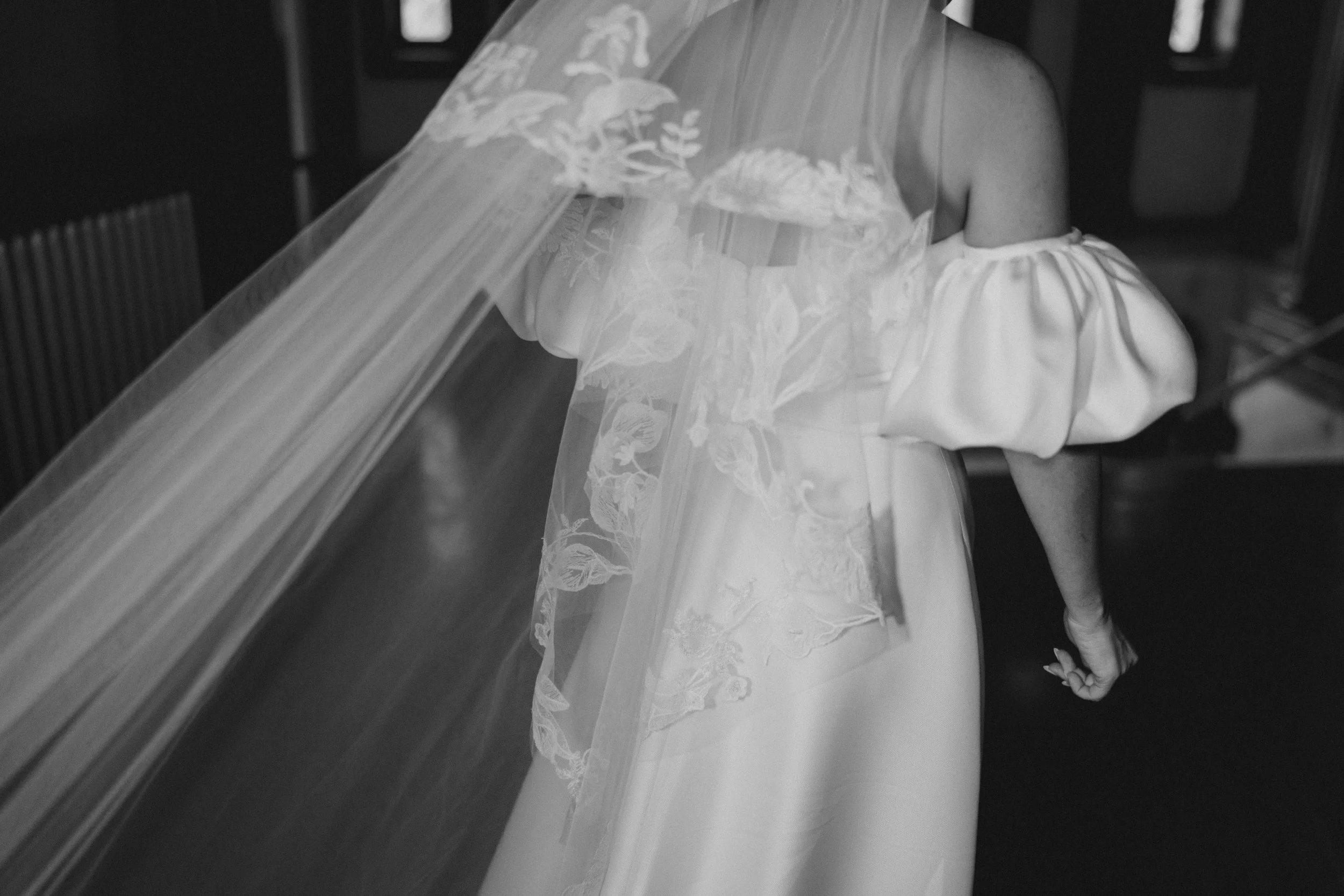 Black and white photo of a woman in a wedding dress with an off-the-shoulder sleeve, semi-transparent veil with floral embroidery, and her hand clenched into a fist.