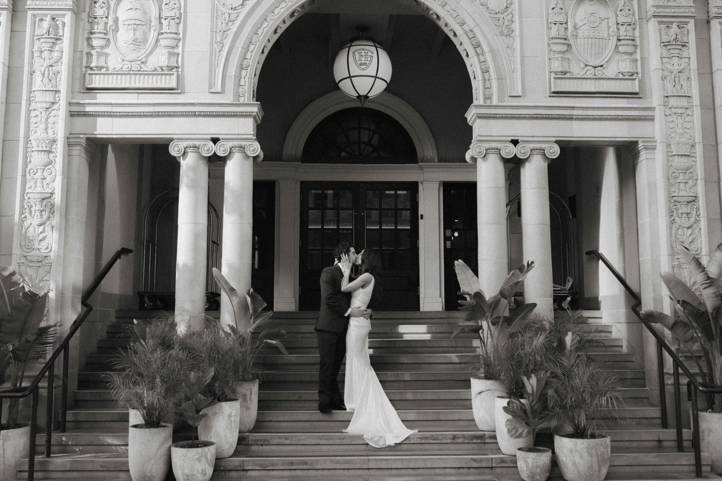 A black and white photo of a couple in wedding attire sharing a romantic moment on the steps of a grand building, surrounded by potted plants.
