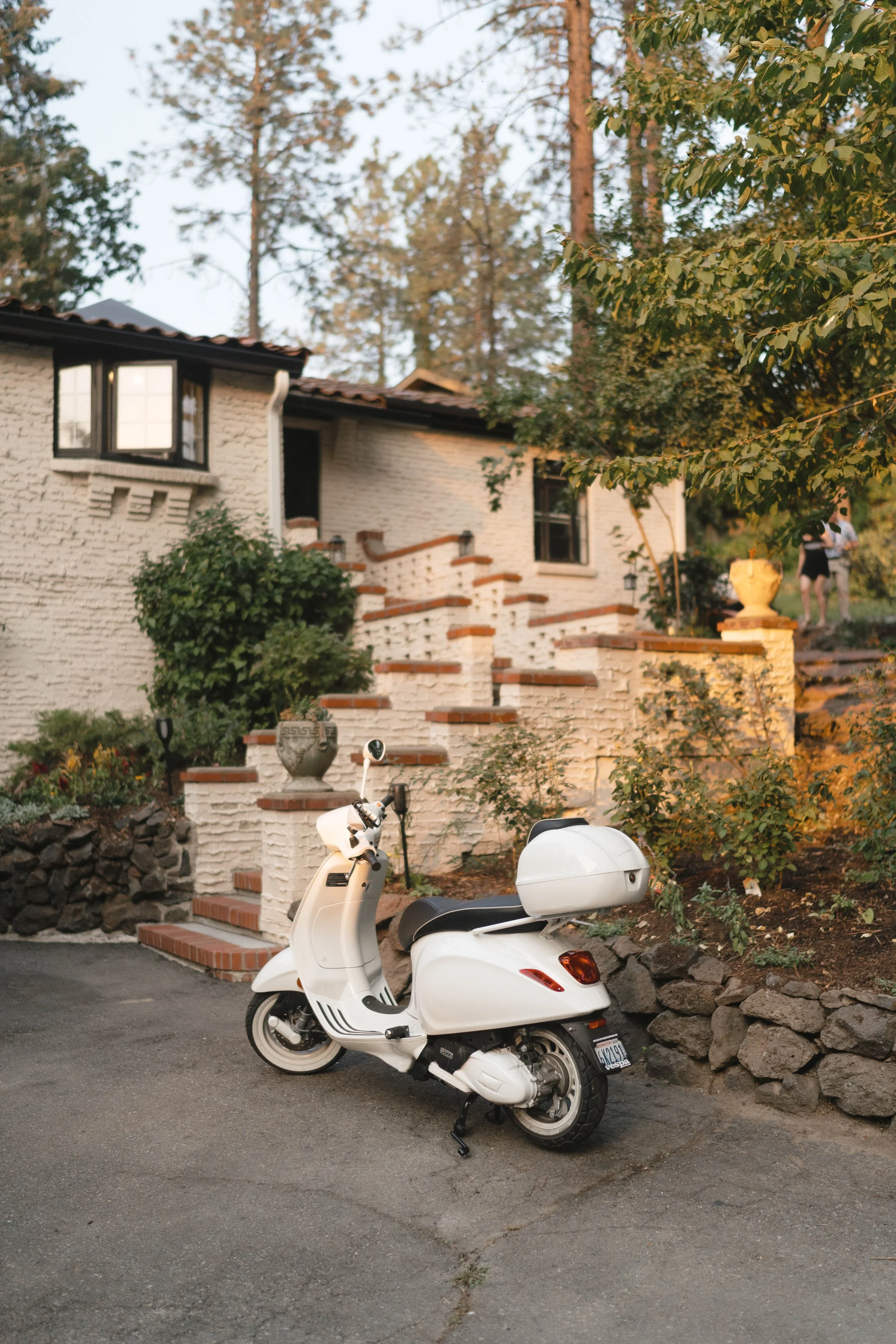 A white scooter parked on a driveway in front of a white brick house with stairs, greenery, and trees in the background.
