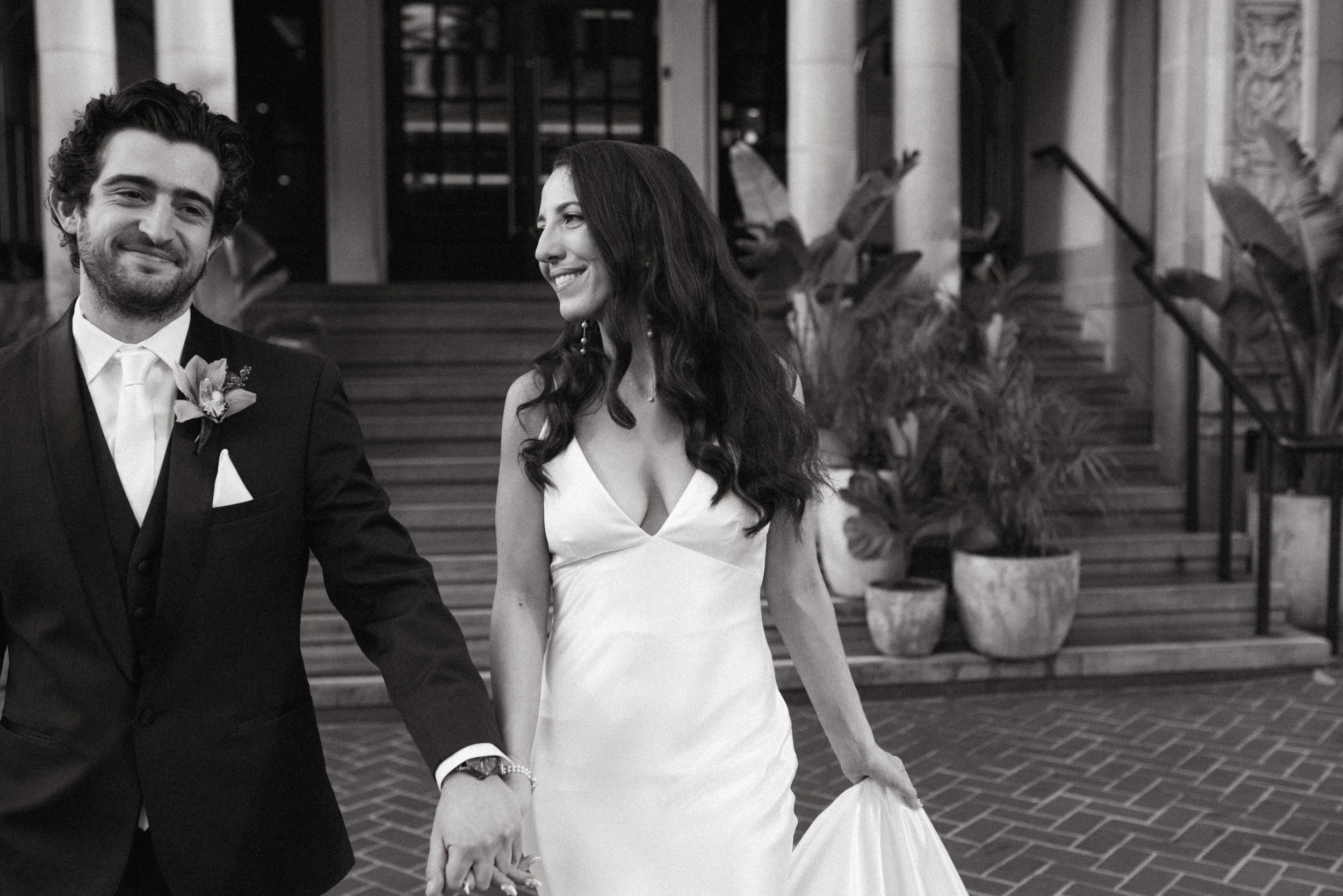 A happy couple in wedding attire holding hands outside a building with stairs and potted plants.