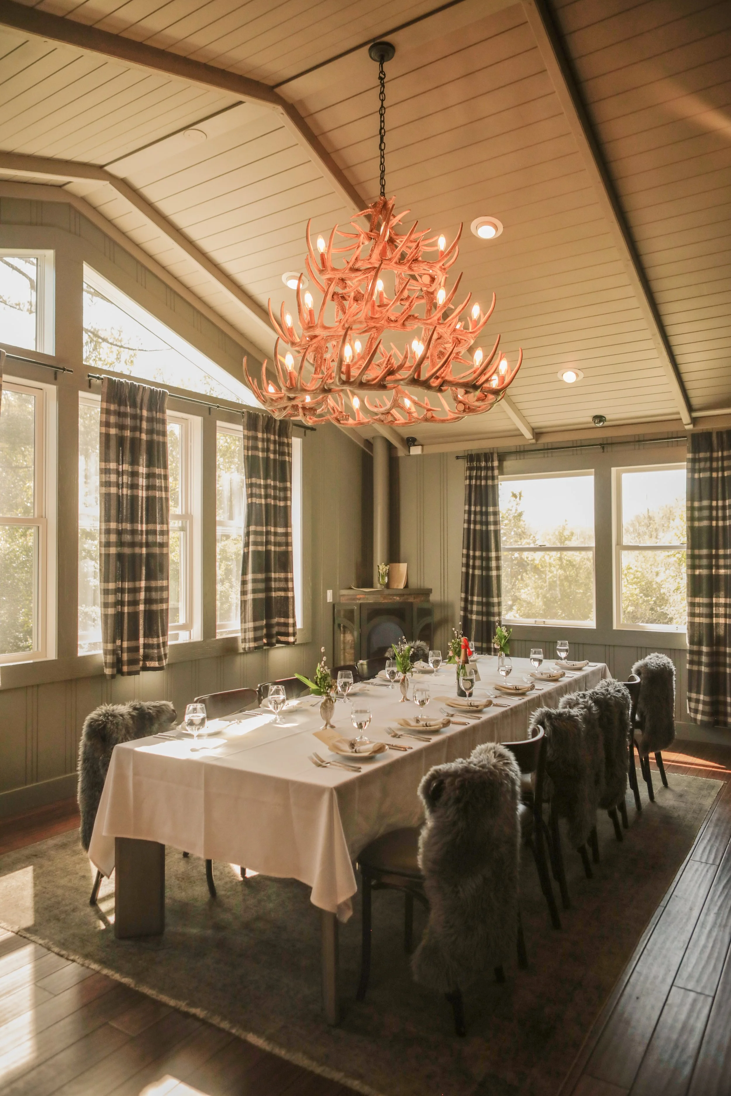 A cozy dining room with a long table set for a meal, surrounded by chairs with faux fur covers, large windows with plaid curtains, and a chandelier made of antlers hanging from the ceiling.