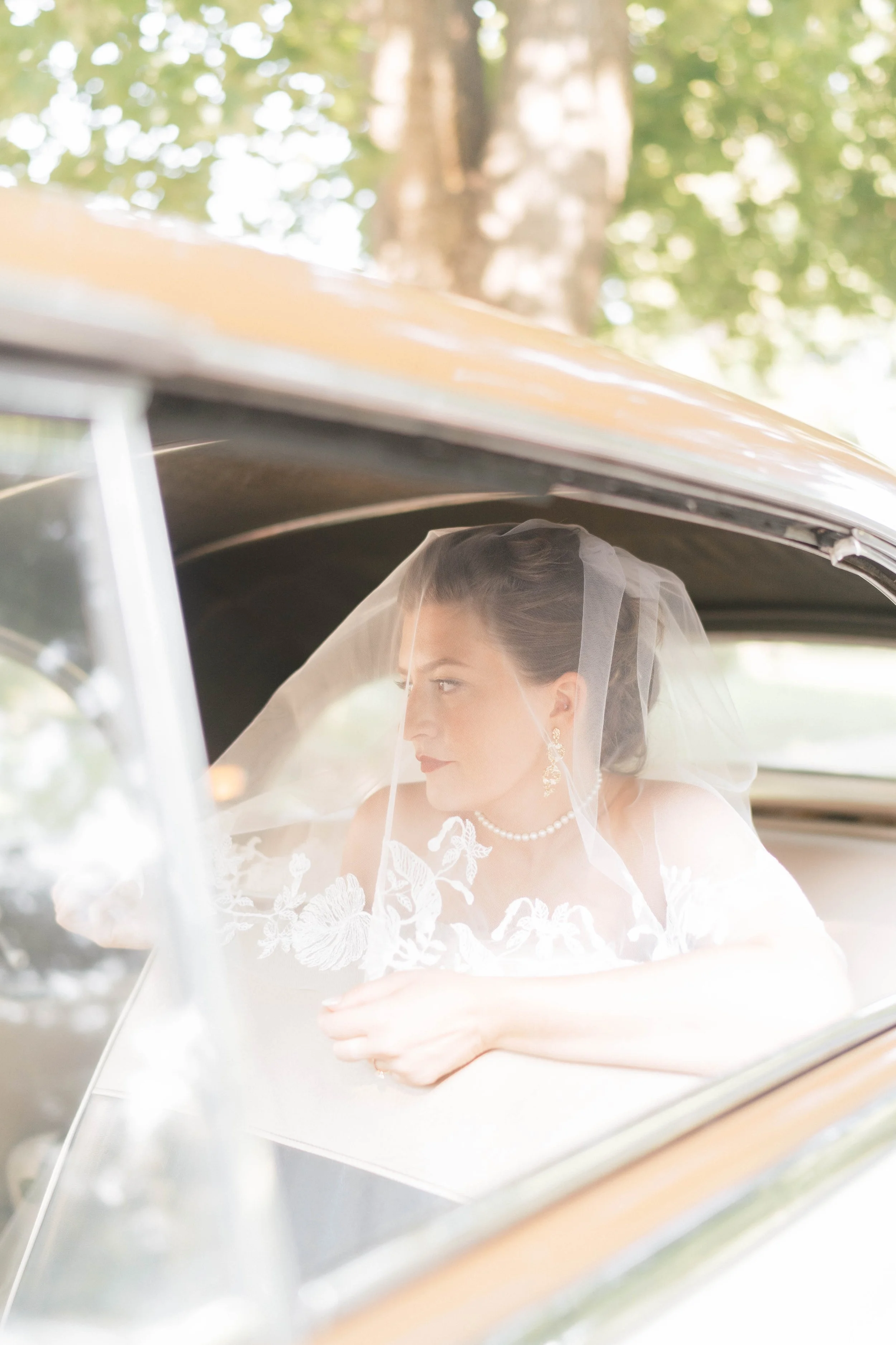 A bride with dark hair, pearl earrings, and a pearl necklace, sitting inside a vintage car with a veil over her face, looking out the window.