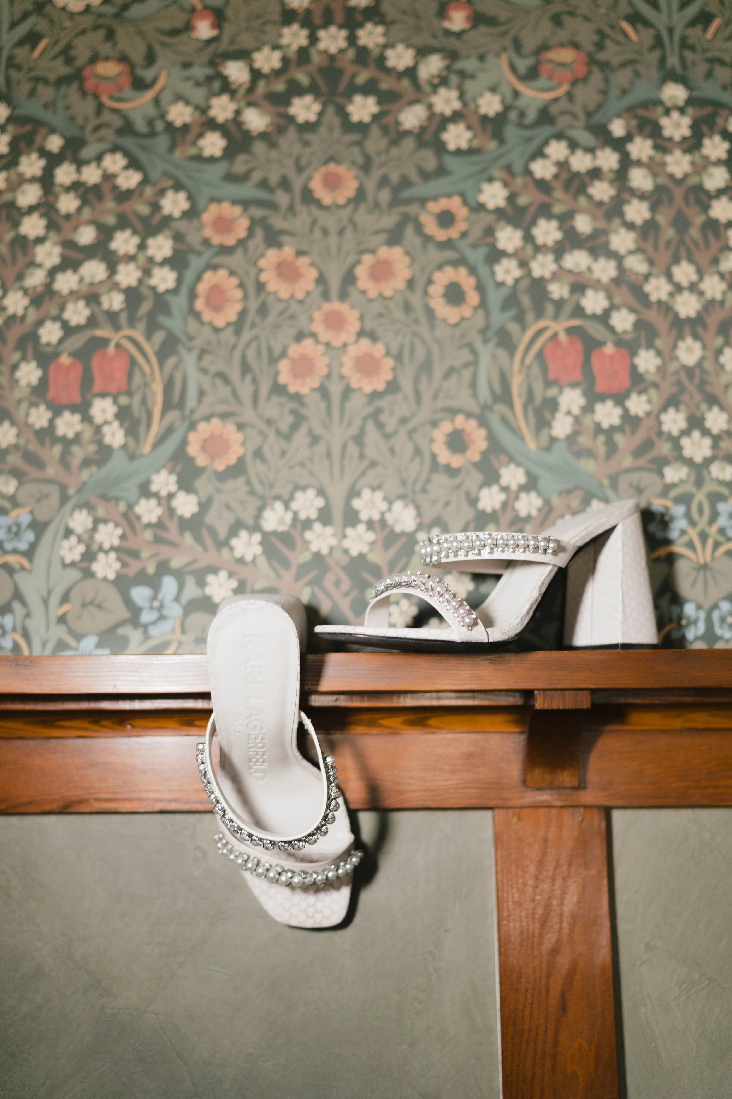 A pair of white high-heeled shoes with rhinestone embellishments placed on a wooden surface against a decorated floral wallpaper background.