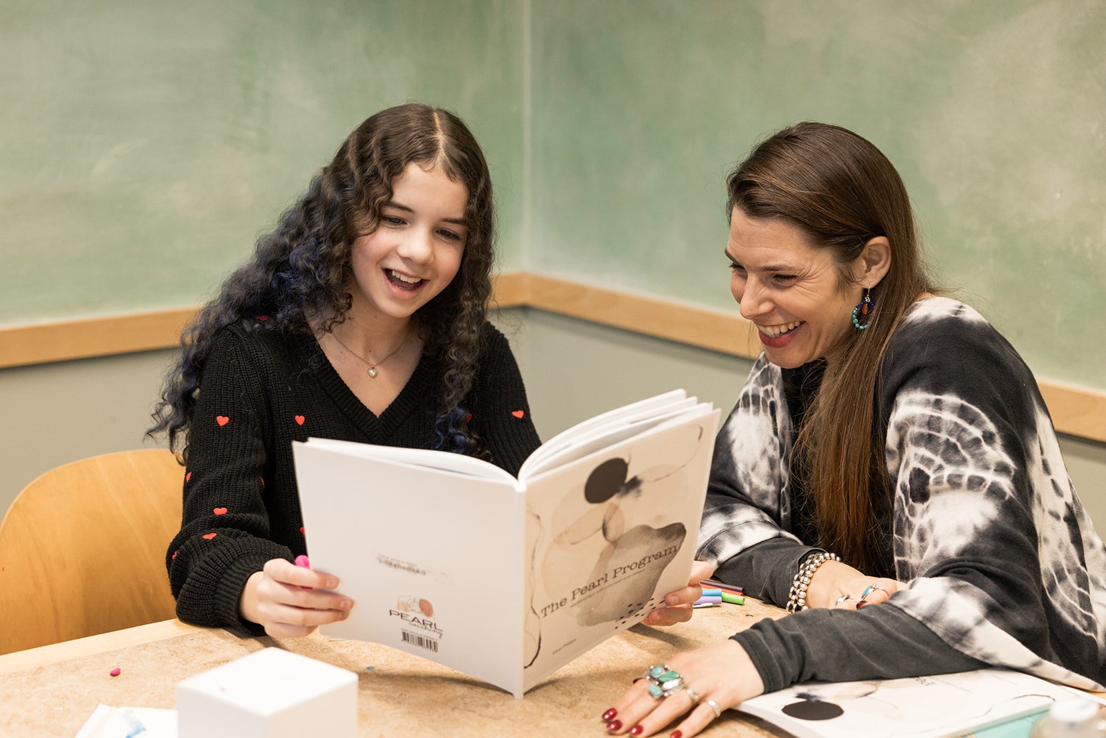 A young girl and an adult woman sitting at a table, looking at a large book and smiling.