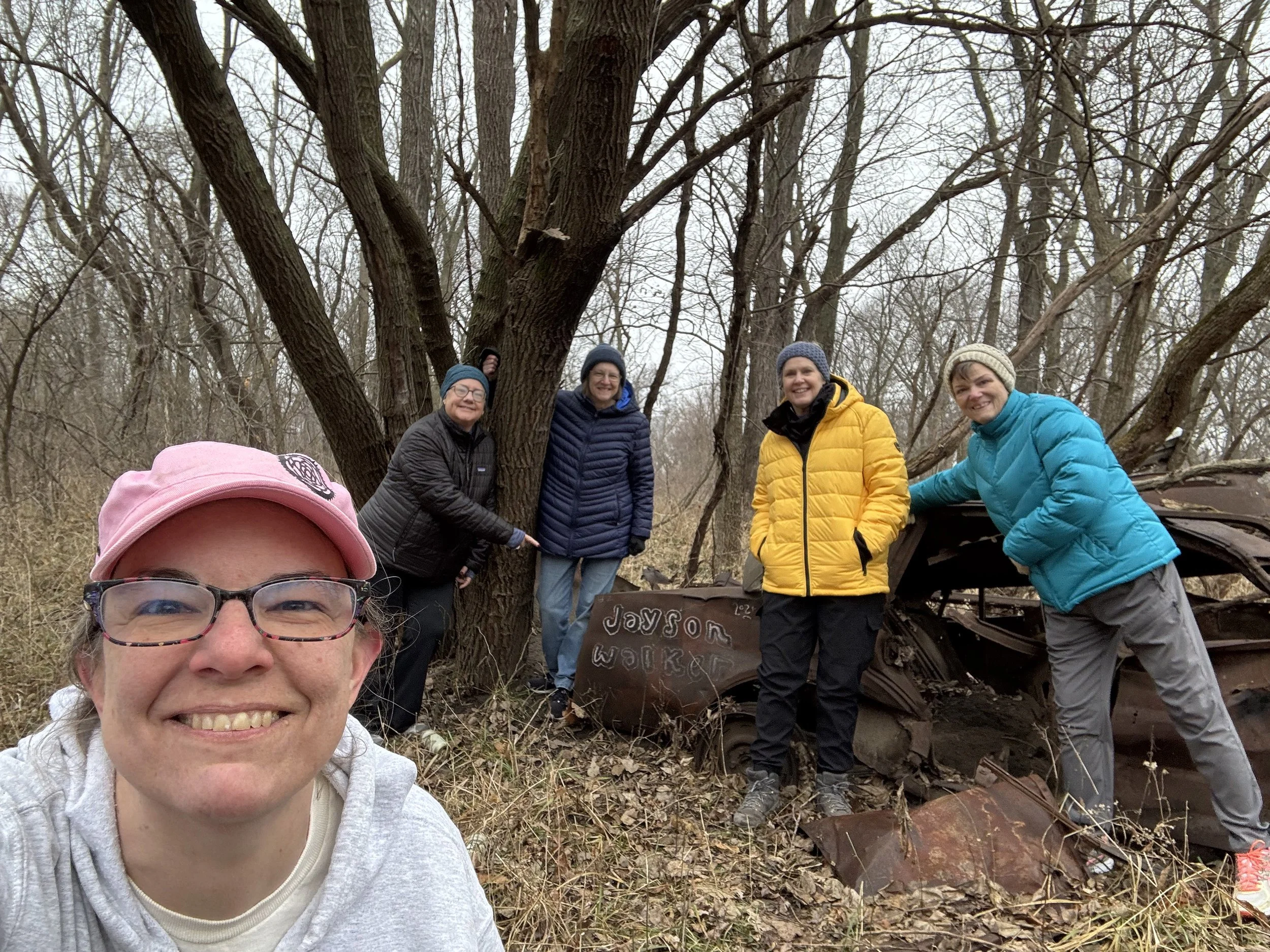 Five hikers pose with a rusted car in the middle of the woods.