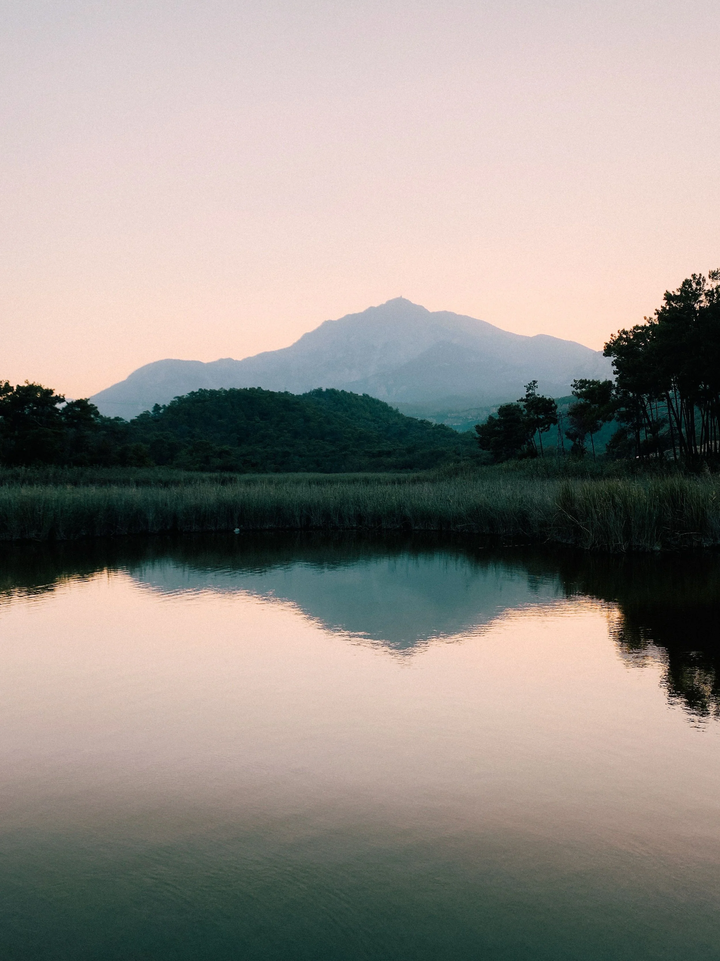 Calm lake with still water reflecting a mountain and trees in the background at dusk or dawn.