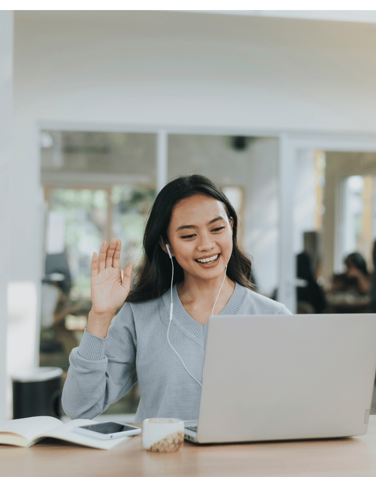 young woman smiling at her laptop screen with earbud headphones, waving at the screen