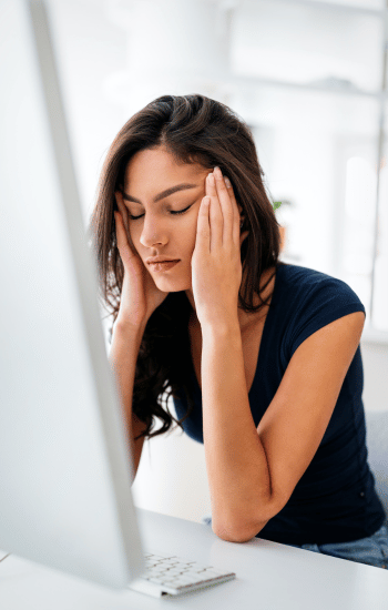 young woman sitting in front of computer screen, holding her temples due to perfectionist frustration