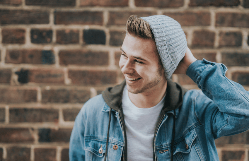 young man in a gray beanie and jean jacket, smiling in front of a brick wall