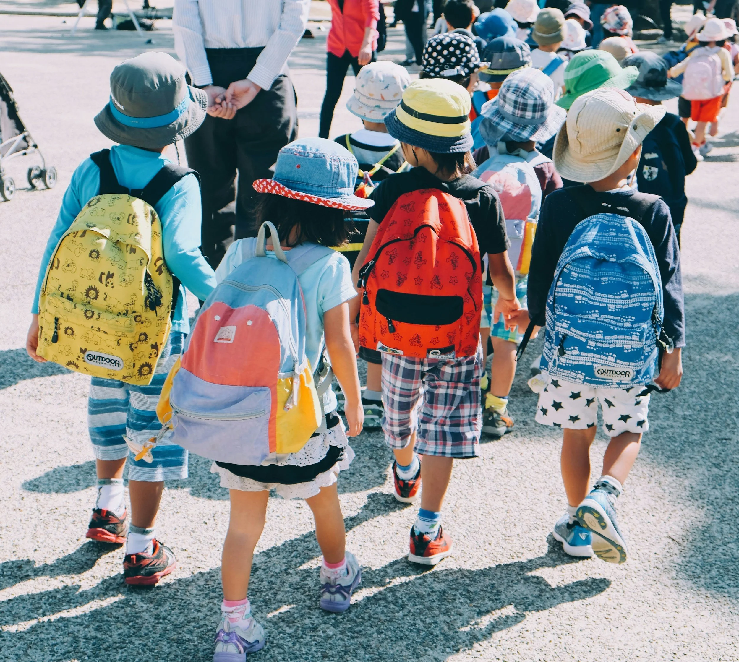 A group of children wearing hats and backpacks walking outdoors on a sunny day, supervised by adults.