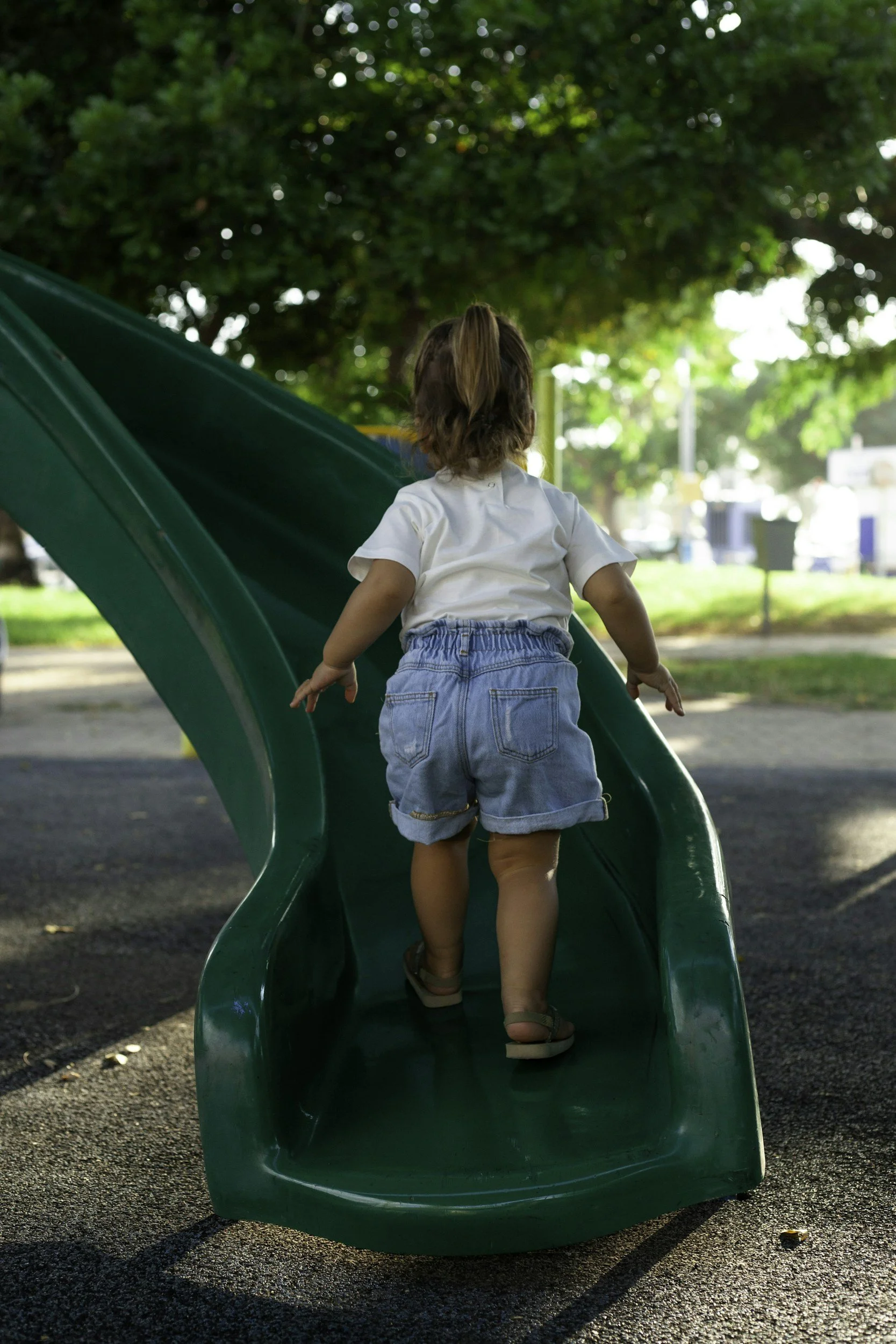 A young girl with brown hair in a ponytail, wearing a white t-shirt and denim shorts, is climbing a green slide in a park during daytime with green trees and park elements in the background.