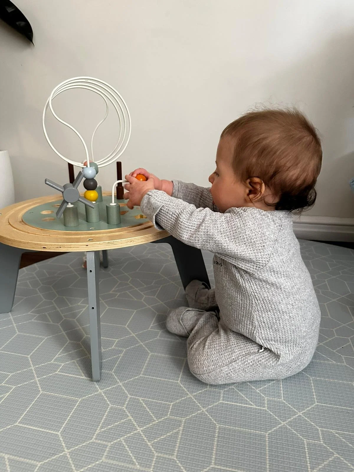 A young child sitting on a grey hexagon patterned rug, playing with a wooden marble run toy at a small table.