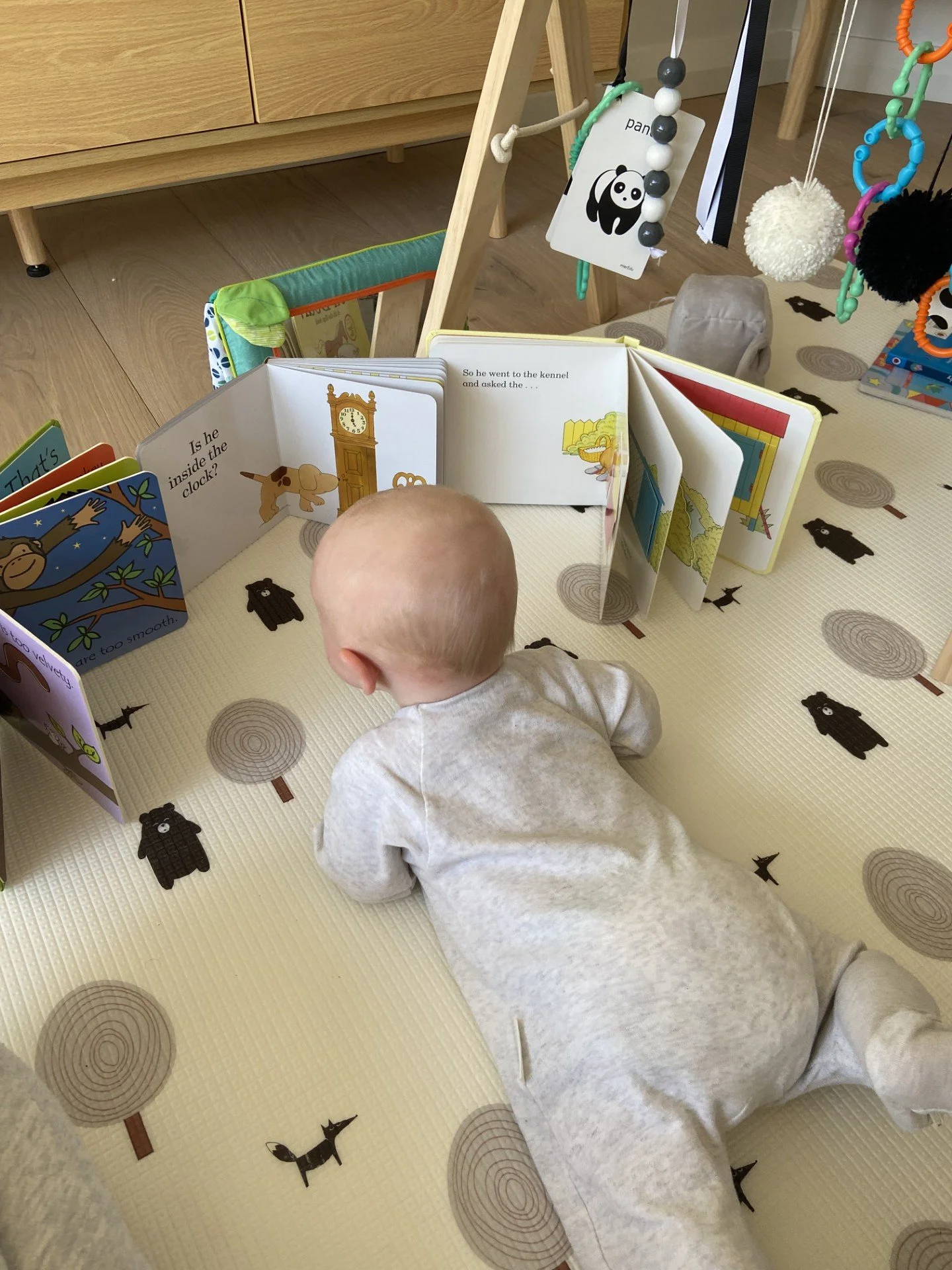 A baby girl lies on her stomach on a play mat with woodland animal illustrations, reading a children's book with colorful pages and pictures of a clock and a dog.