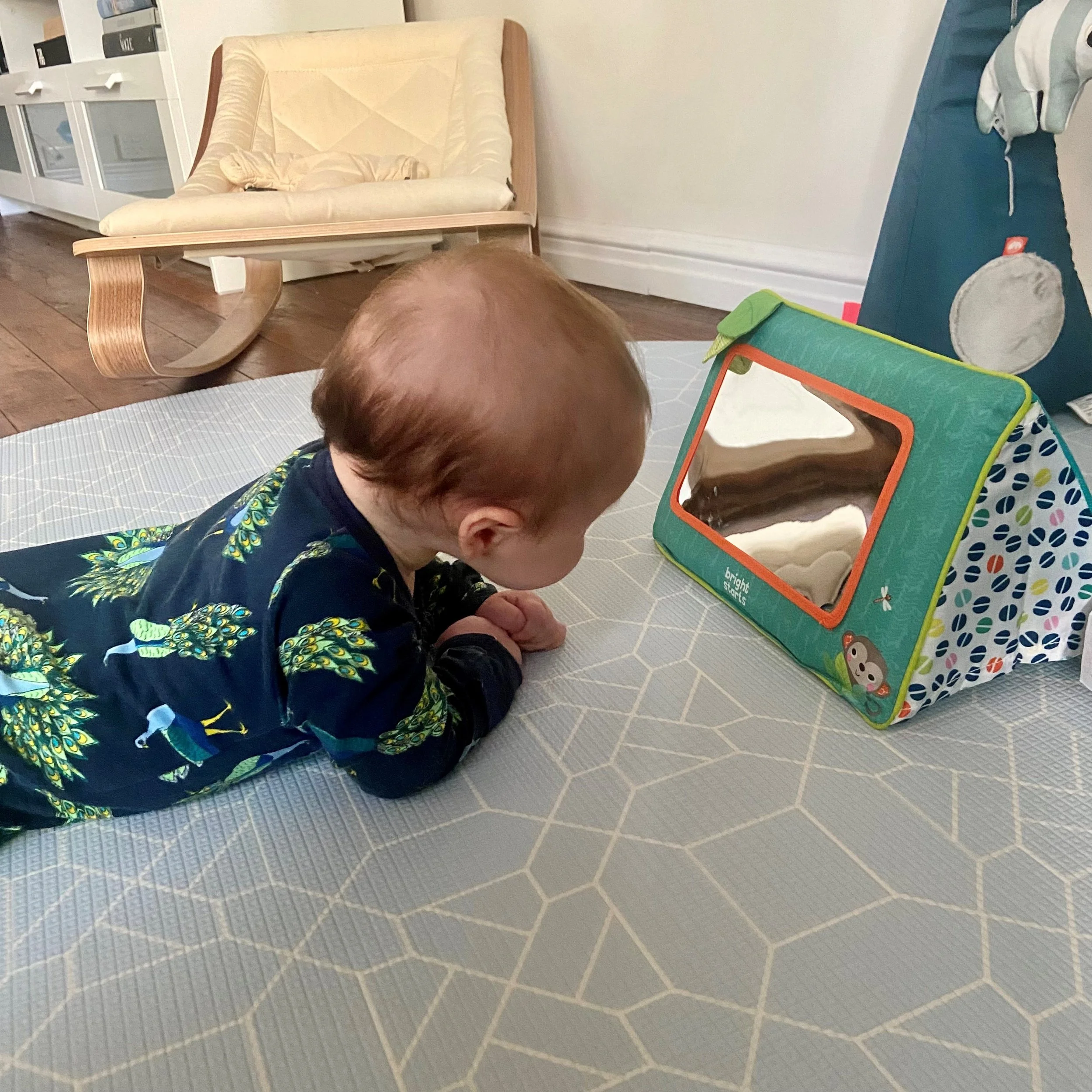 A baby with light brown hair and wearing a dark pajamas with colorful dragon patterns, lying on a geometric patterned mat, looking towards a small portable mirror on a table.
