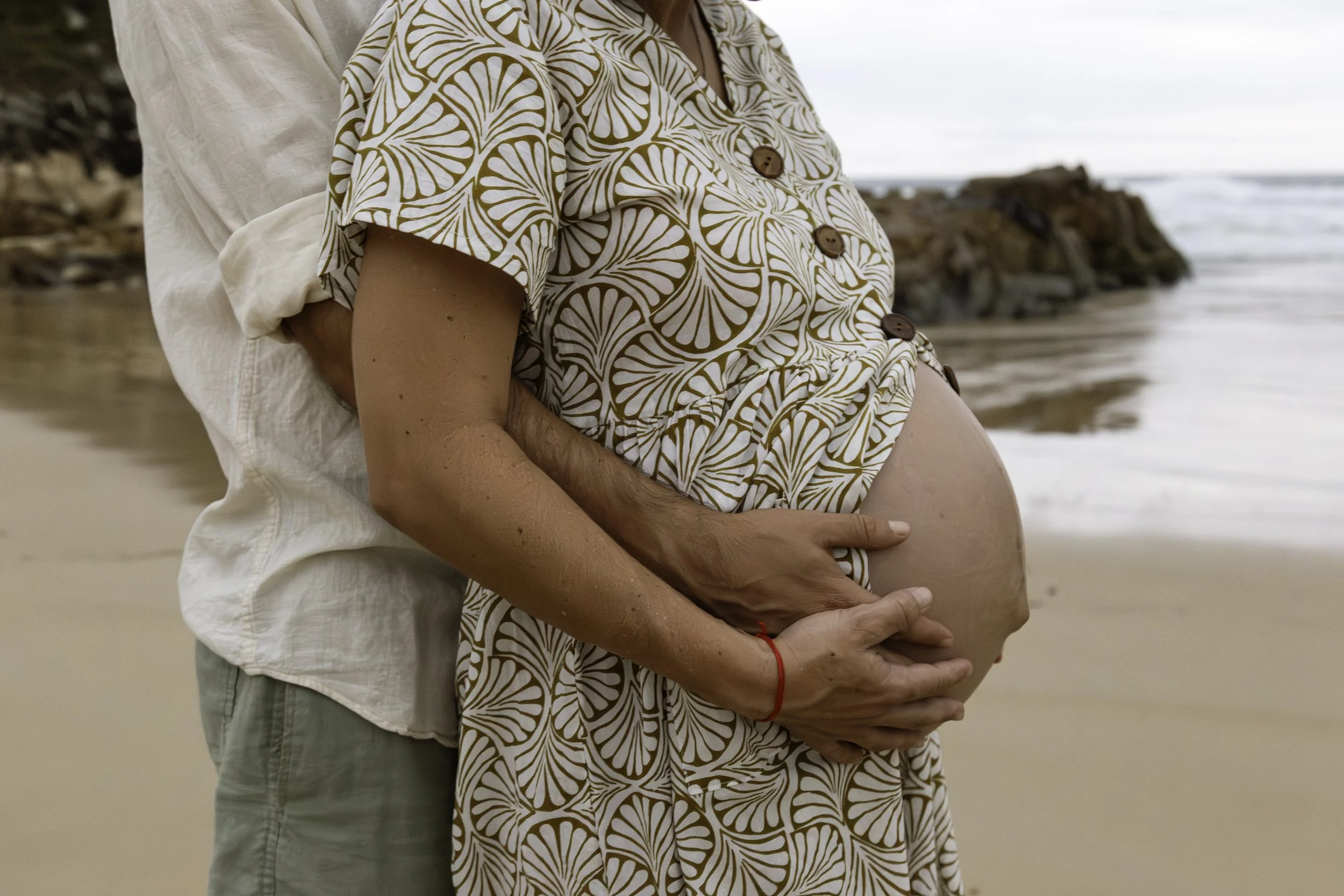 A pregnant woman and a man standing on the beach, with the man holding her belly from behind. The woman is wearing a patterned dress, and the man is wearing a light-colored shirt. The background shows the shoreline and a rocky formation by the water.