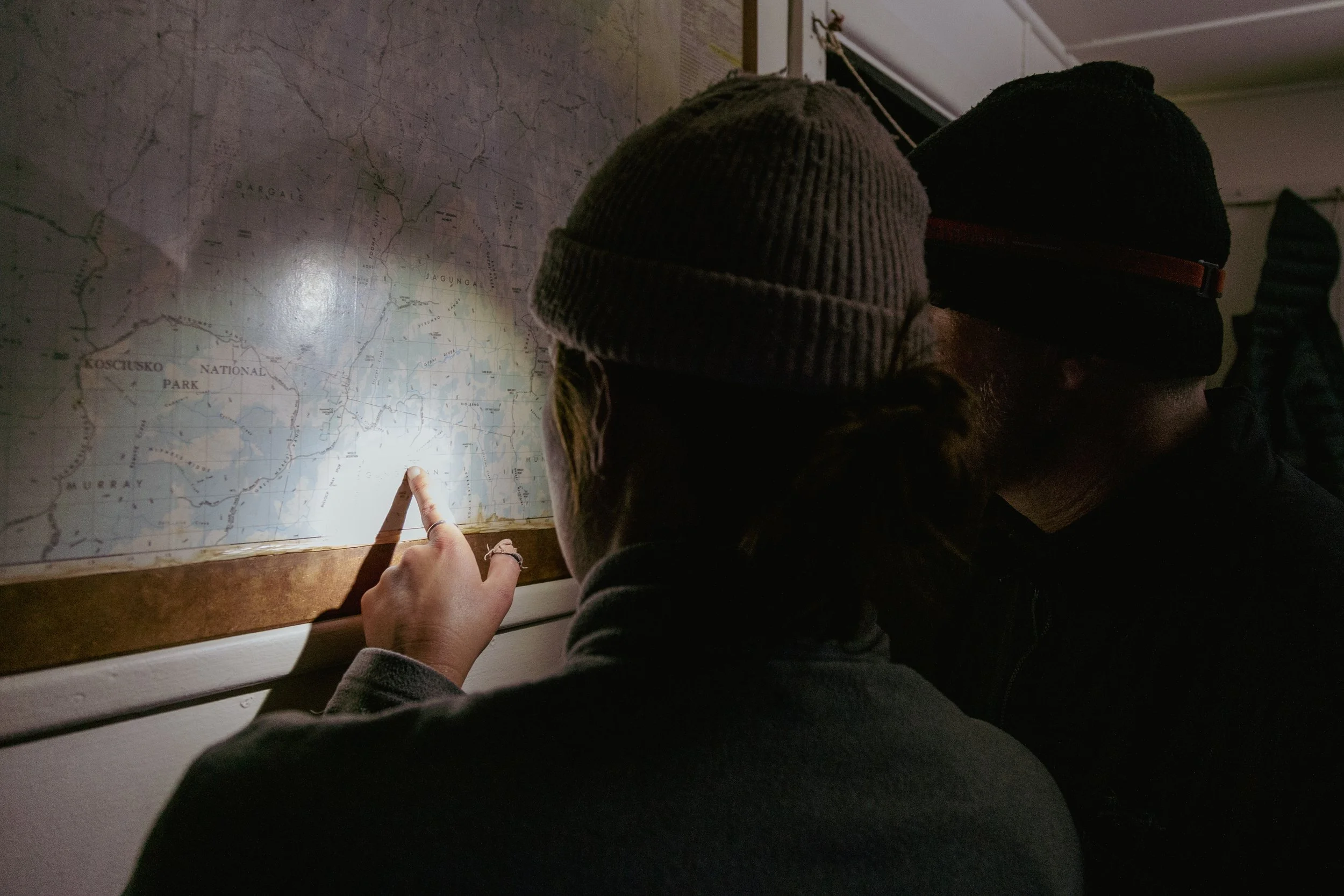 Two people wearing beanies look at a map on a wall, with one person pointing at a location under torchlight.