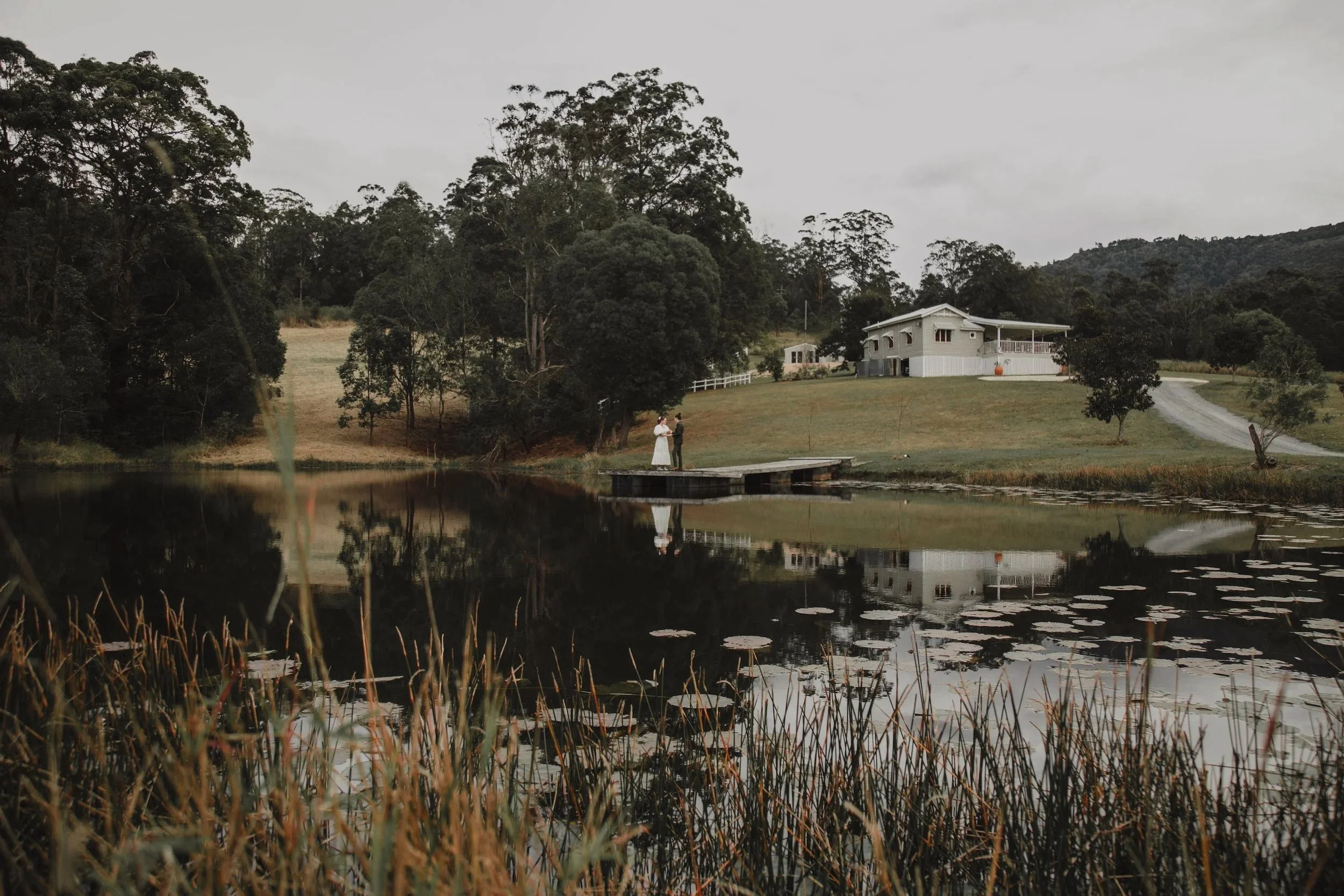 A couple on a small dock by a pond with lilies, surrounded by trees and a white house on a hill in the background.