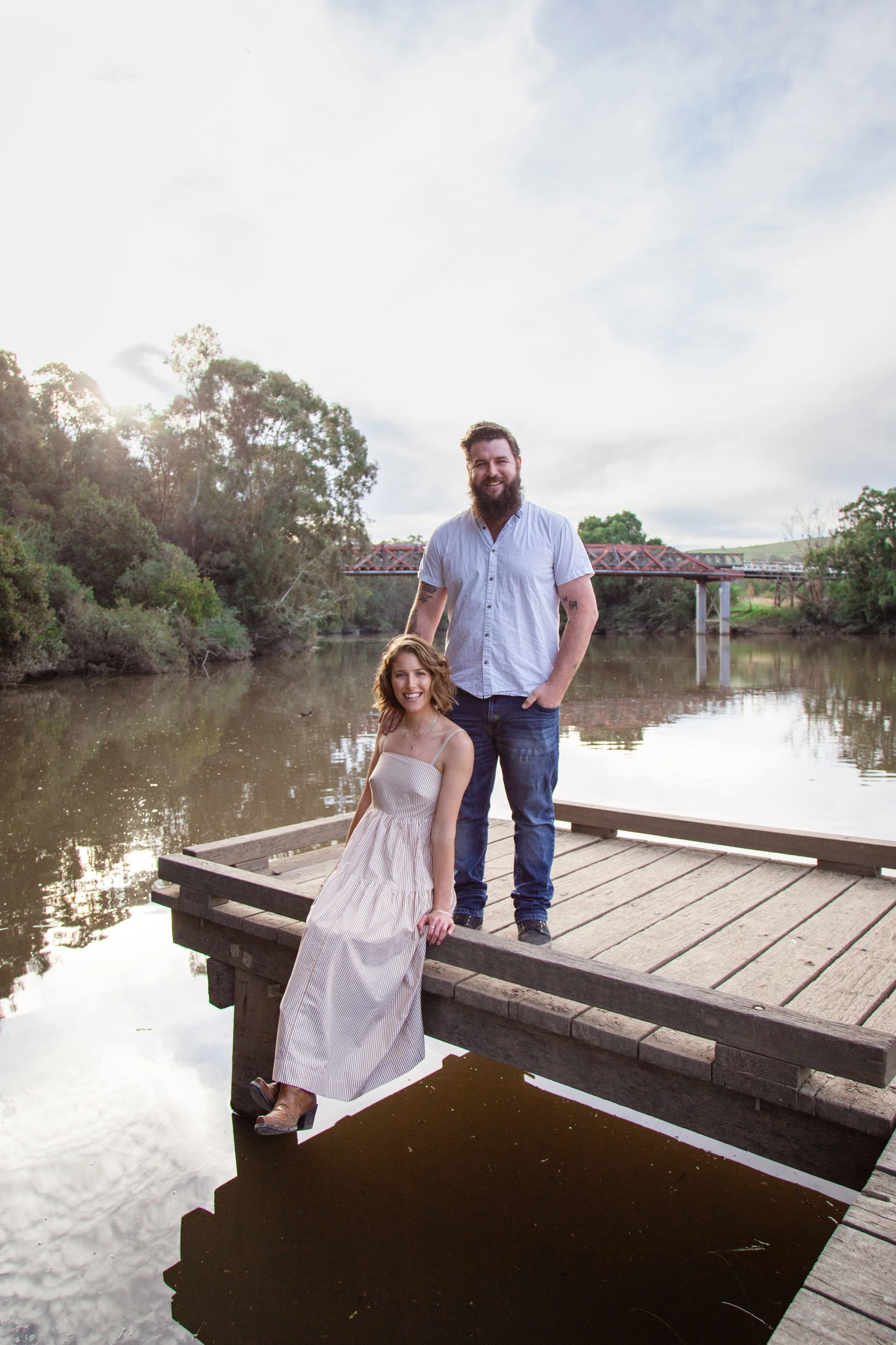 A man and a woman posing on a wooden dock by a river, with trees and a bridge in the background, during daylight.