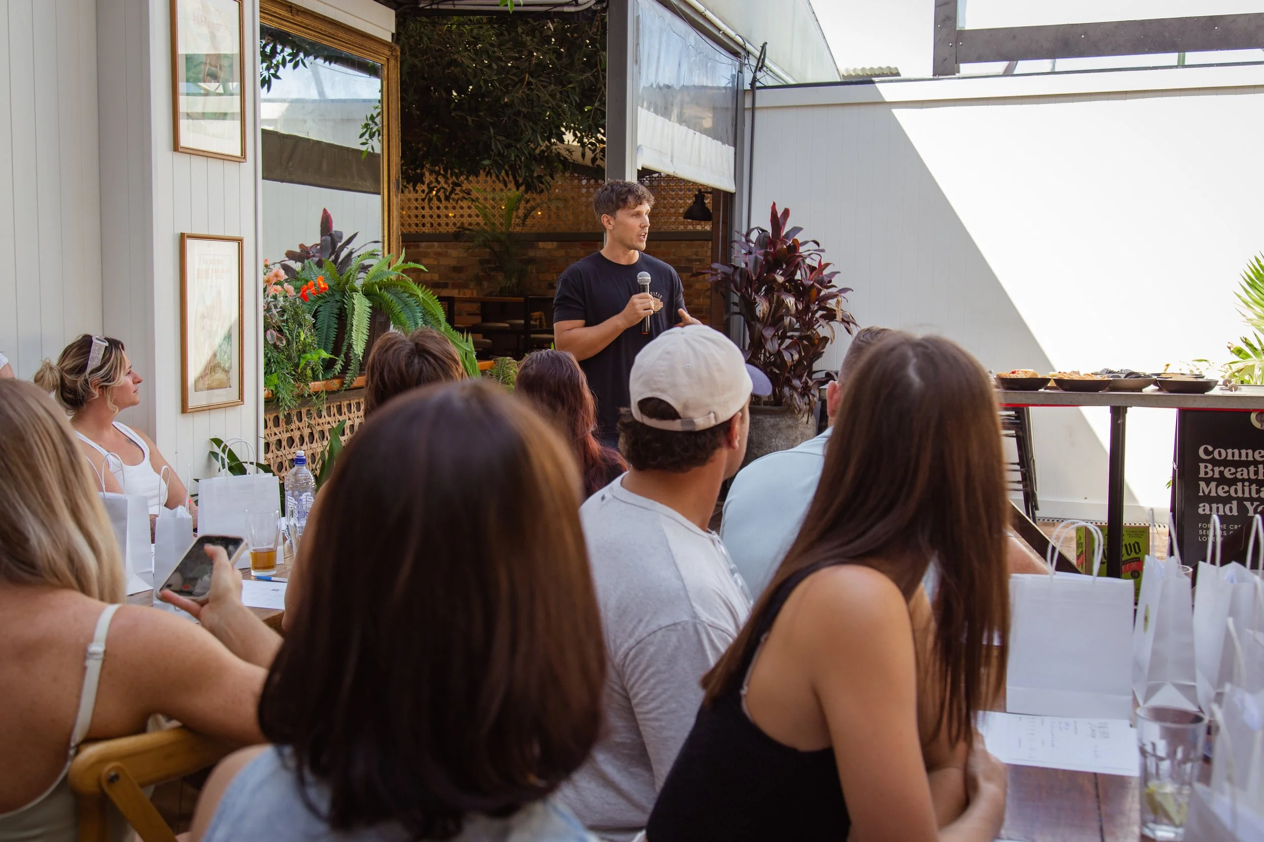 A man speaking into a microphone in front of an audience at an indoor event space with plants and artwork on the walls.