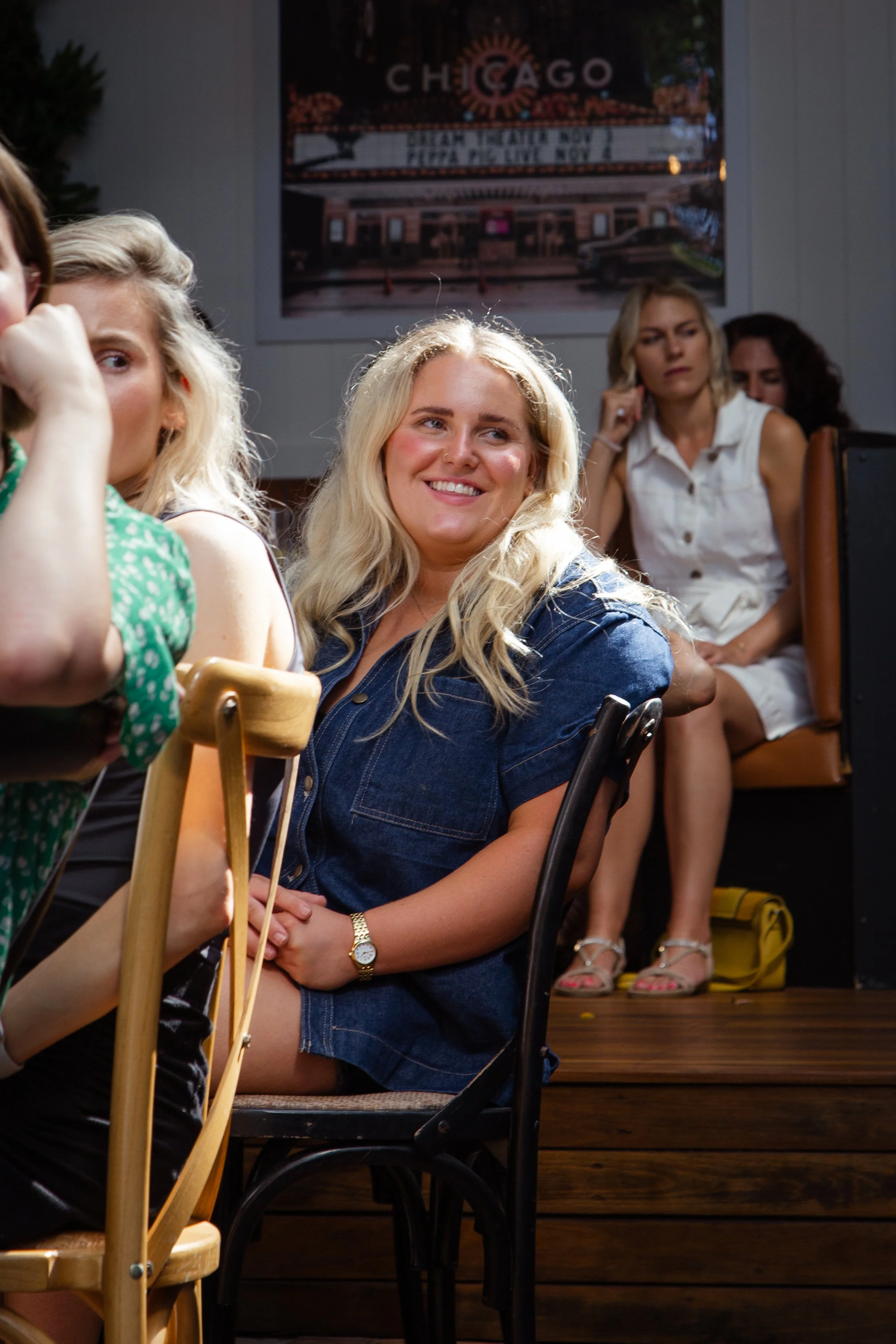 A group of women sitting at an event in a beer garden, with a young woman smiling in the centre, a sign on the wall behind them, and some women looking off to the side.