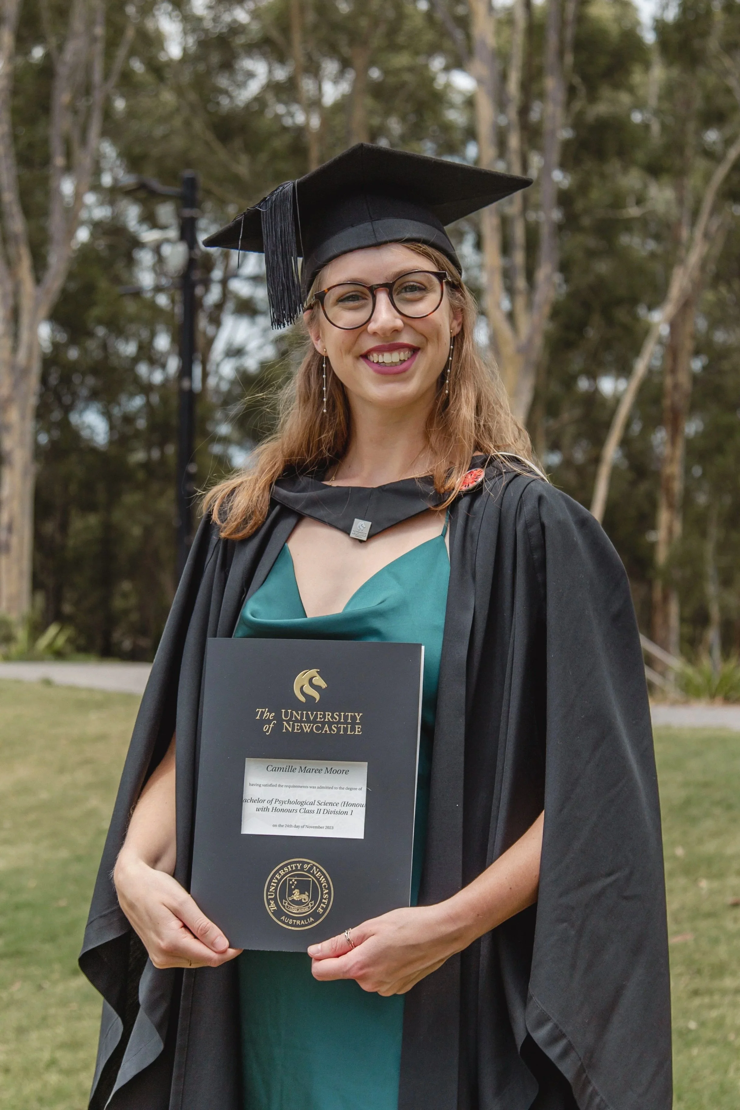 A young woman beams proudly in a graduation cap and gown holding a university certificate outdoors.