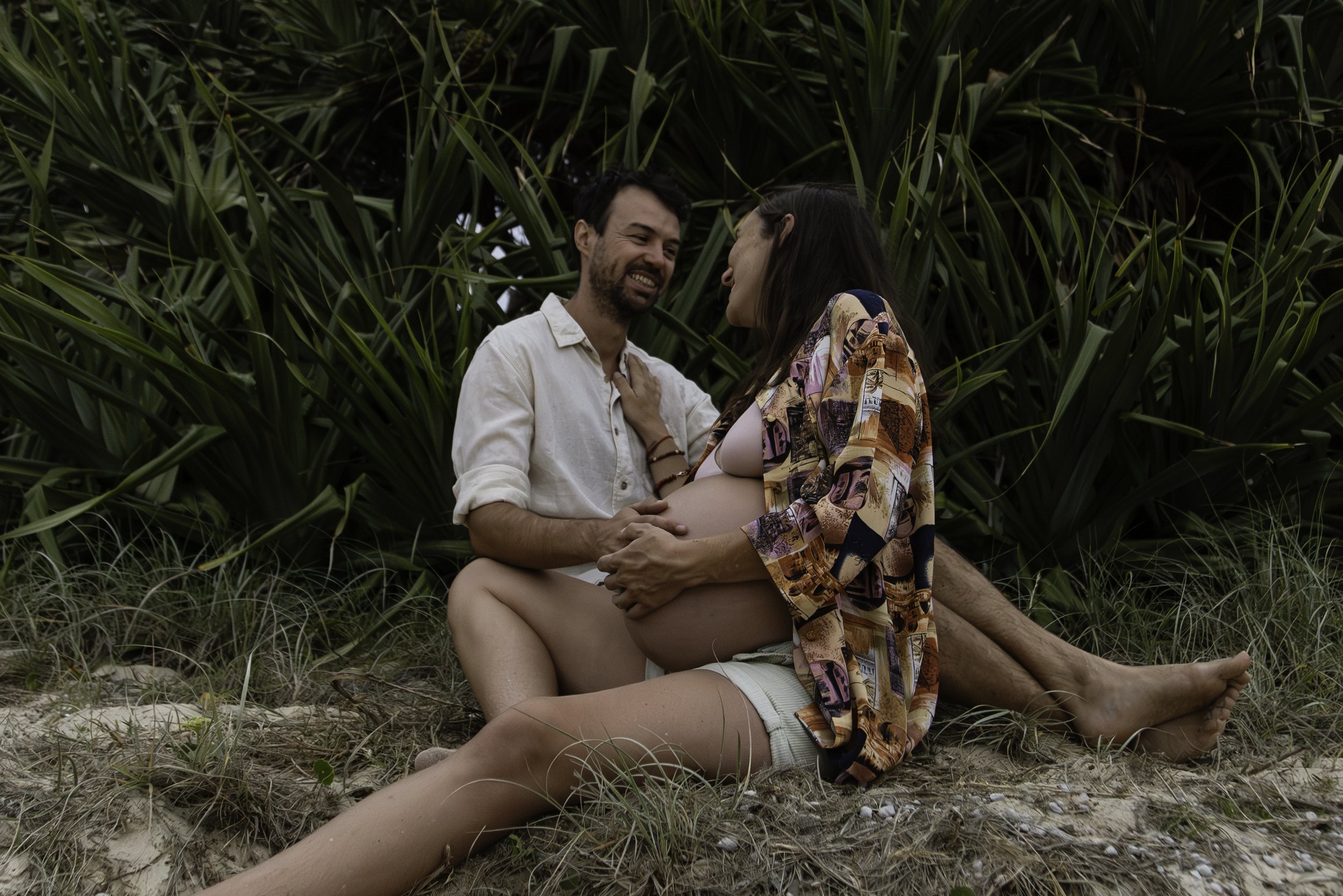 A pregnant woman and a man sitting on the sand near large green plants, smiling and looking at each other.