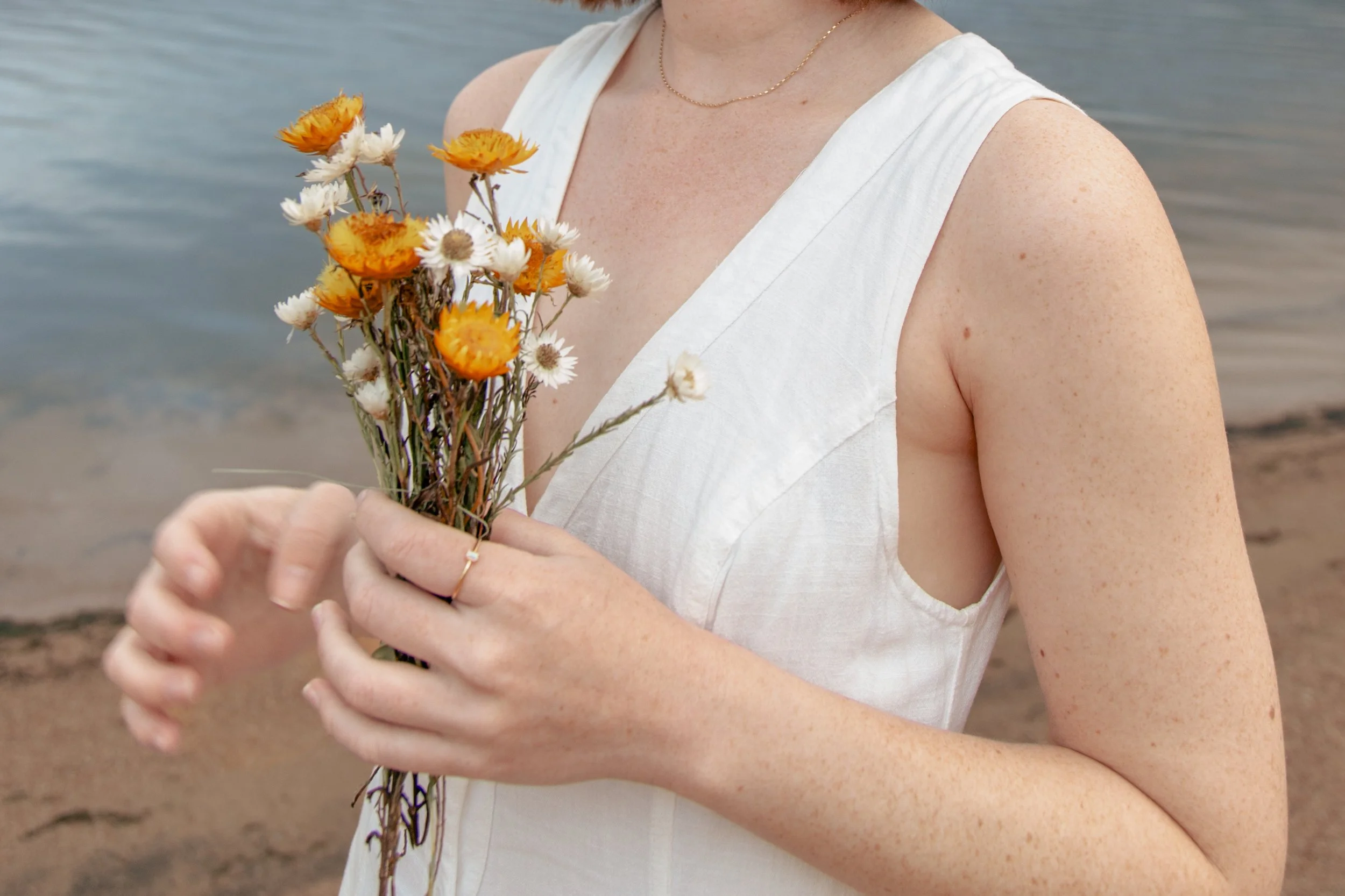 A woman in a white sleeveless dress holding a bouquet of orange and white flowers by a lakeshore.