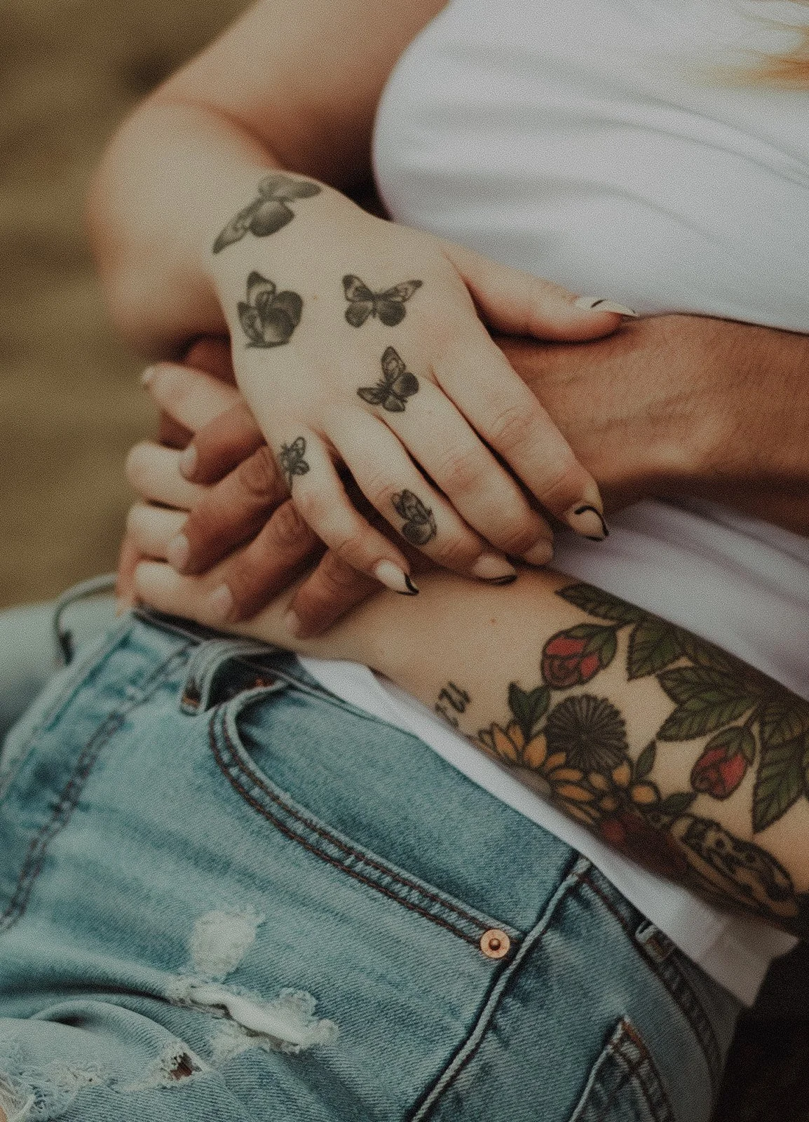 Close-up of hands with tattoos, including butterflies on one hand and flowers on the arm, resting on denim shorts and a white top.